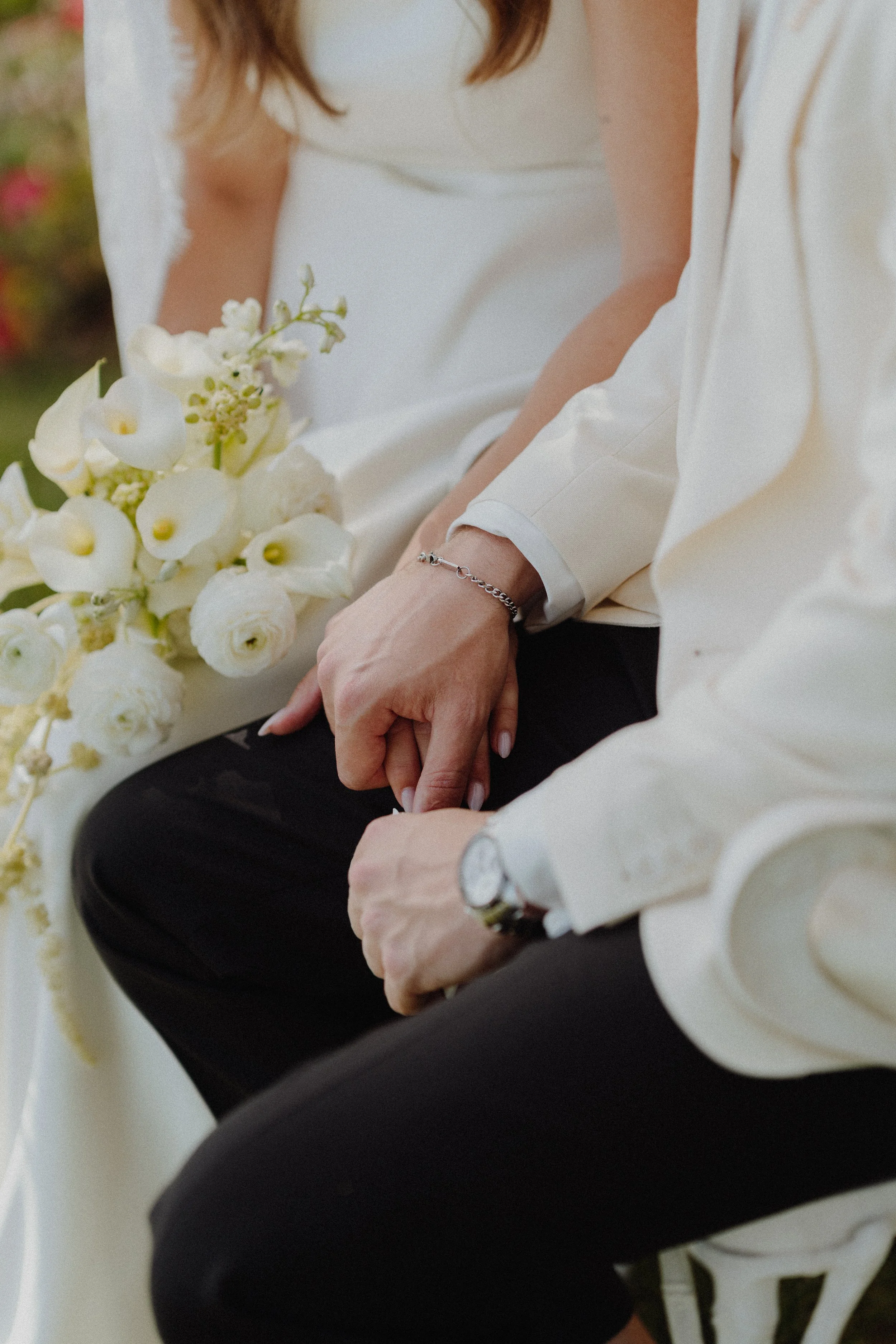 Close-up of a woman sitting on a white chair, holding a bouquet of white flowers, dressed in formal attire including a white shirt and black pants, wearing a silver bracelet and wristwatch.