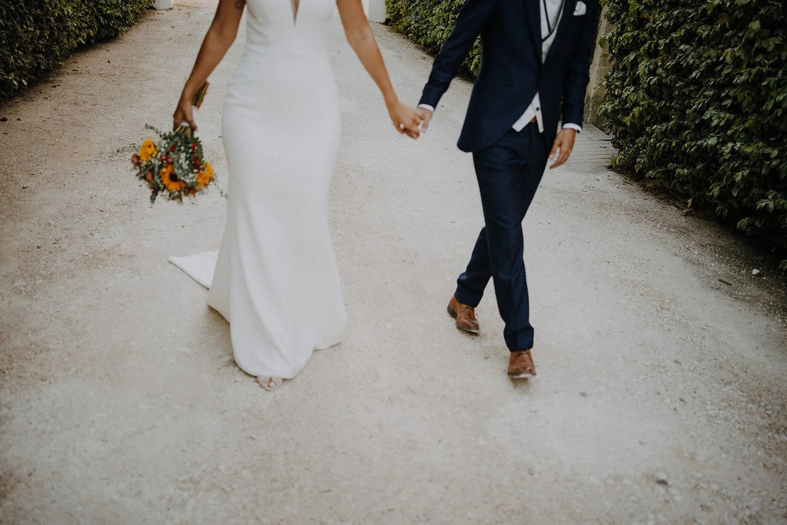 A bride and groom walking hand in hand on a gravel path surrounded by greenery, the bride holding a bouquet of flowers.