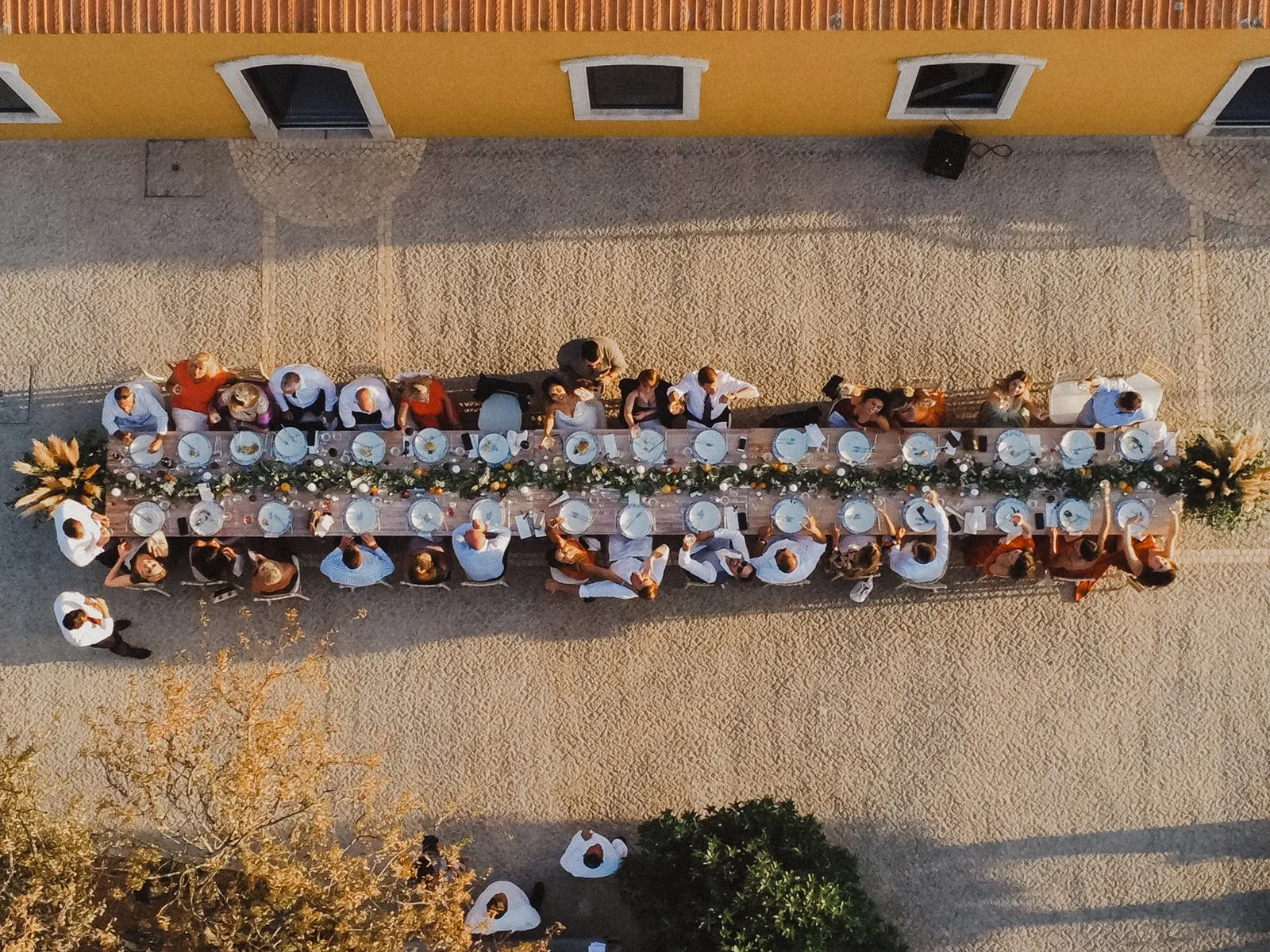 Overhead view of a long outdoor dining table with people celebrating a gathering, set with white plates, silverware, and floral centerpieces, next to a yellow building with windows.