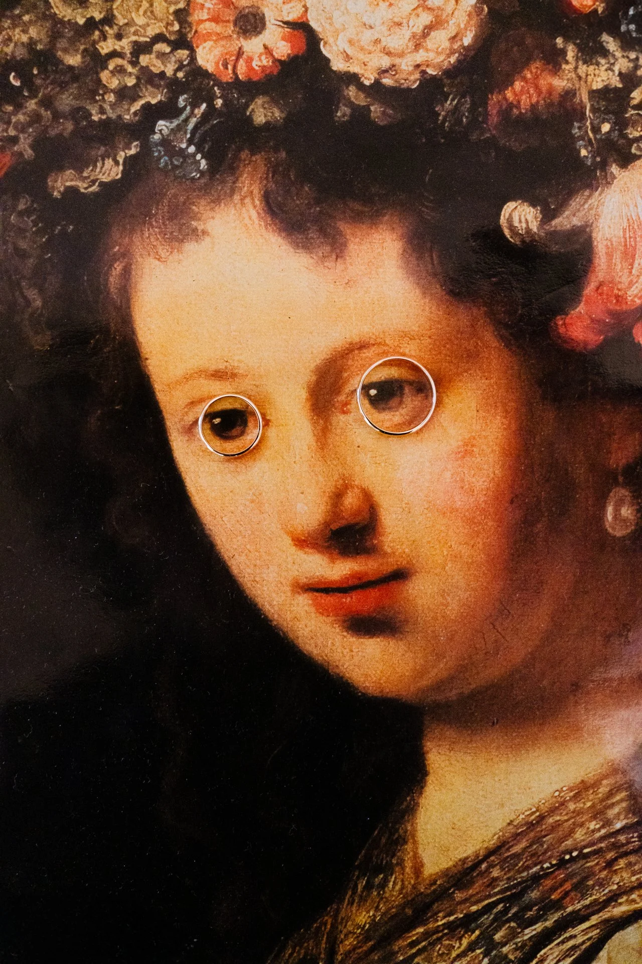Close-up of a framed classic portrait of a woman with curly hair, adorned with a floral crown; a mirror reflection shows the woman's eyes.