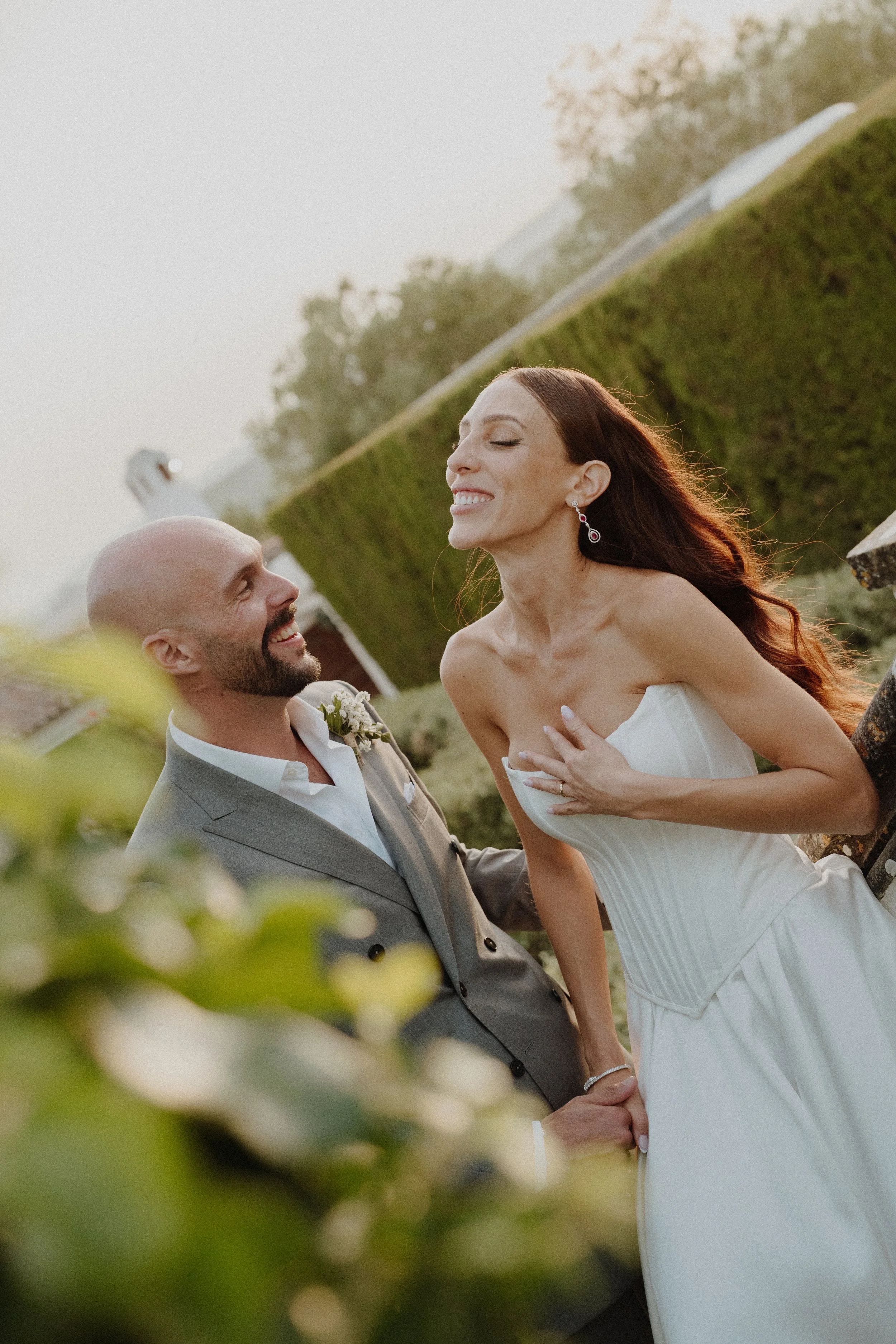 A bride and groom sharing a joyful moment outdoors, holding hands, with the bride smiling and the groom looking up at her, surrounded by greenery.