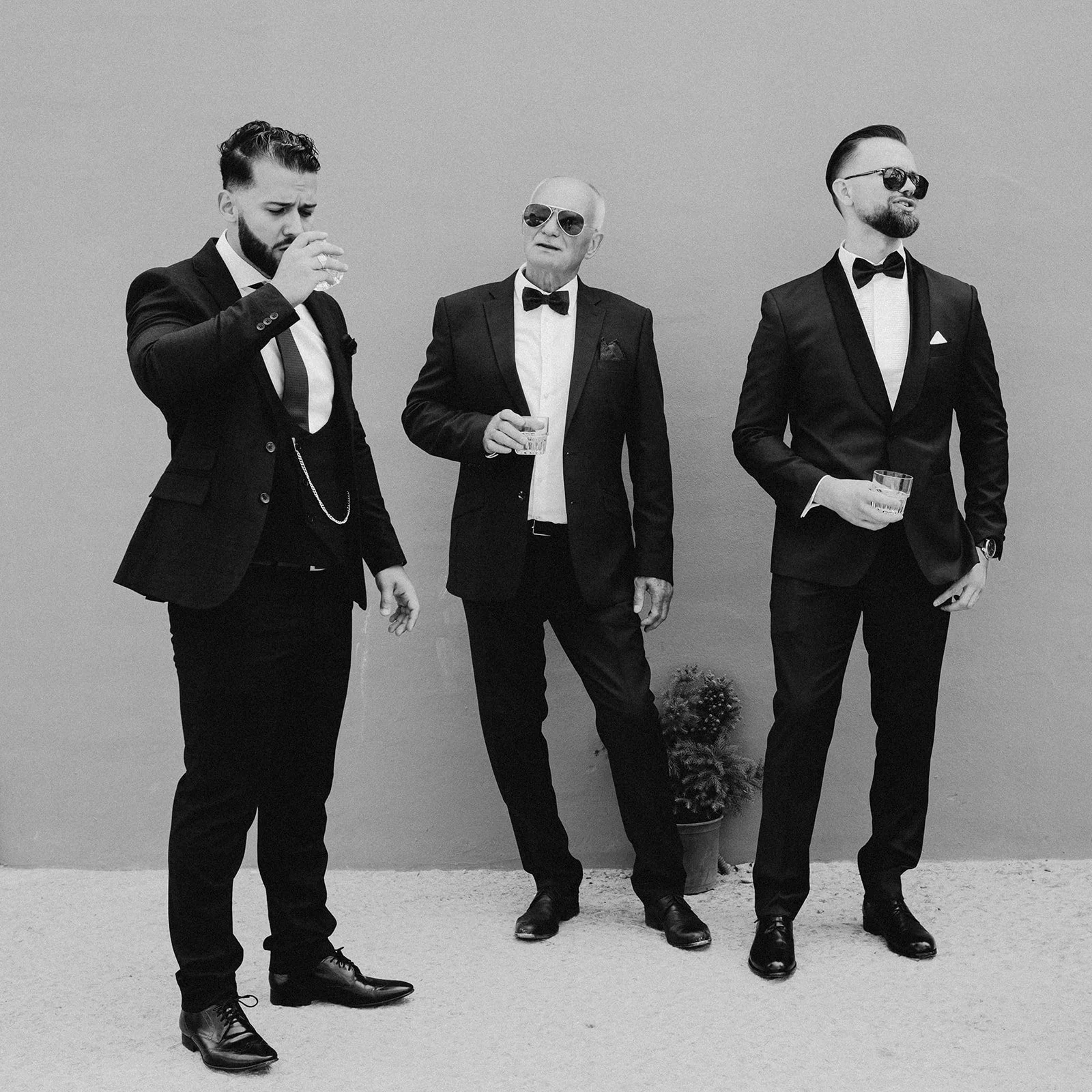 Three men in tuxedos standing against a plain wall, holding drinks, with a potted plant on the ground behind them, black and white photograph.
