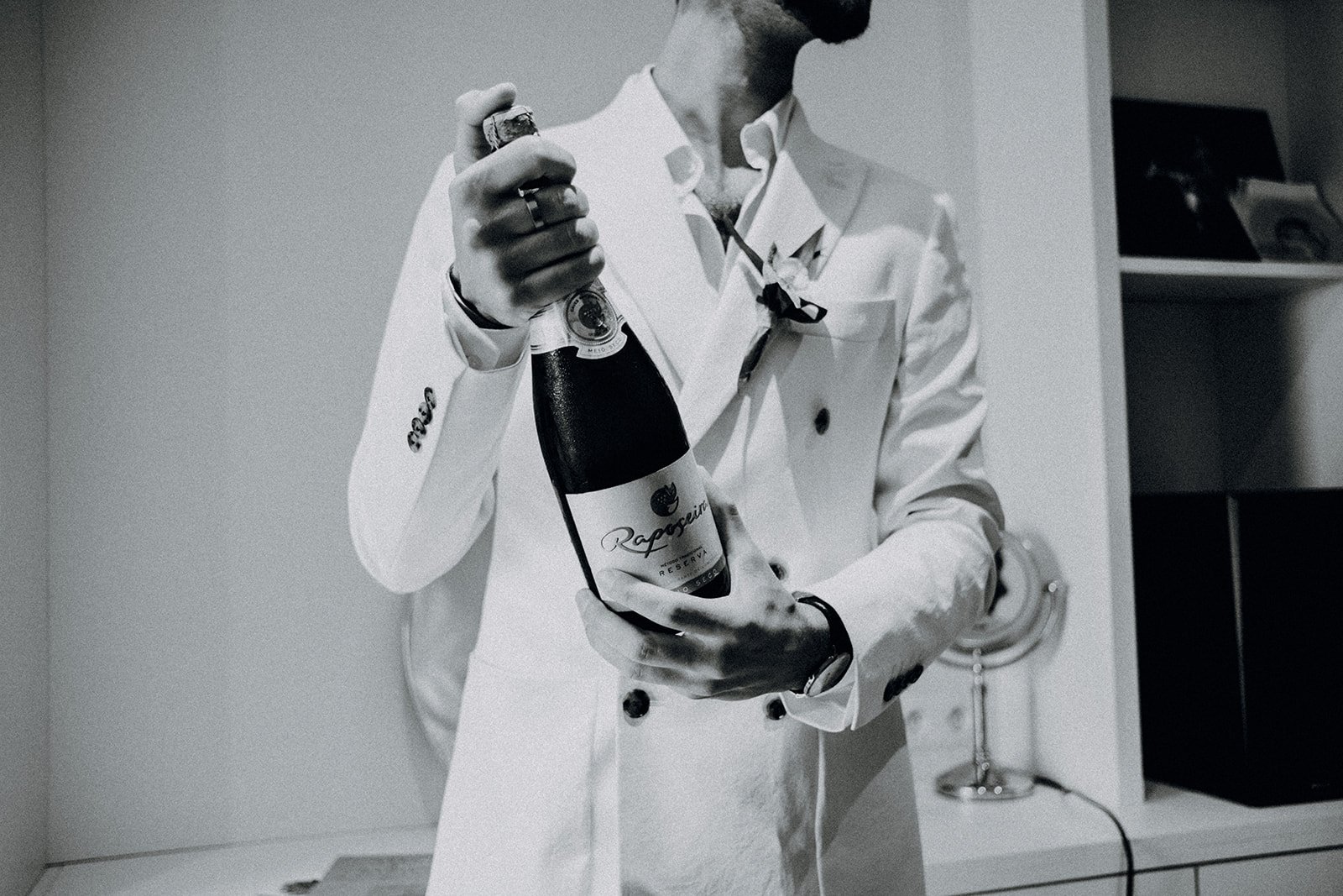 Man in a white suit holding a bottle of champagne in a room with a fireplace and shelf.