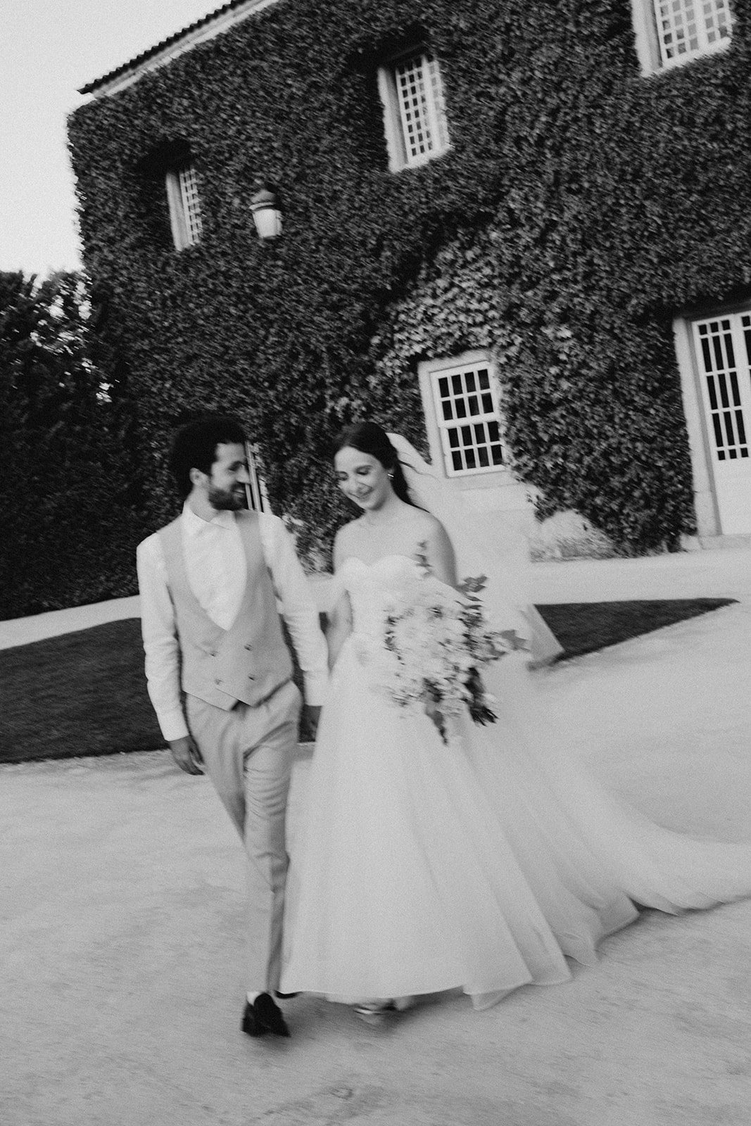 Black and white photo of a bride and groom walking together, smiling, outside a building covered in ivy, with the bride holding a bouquet. Solar de Pancas.