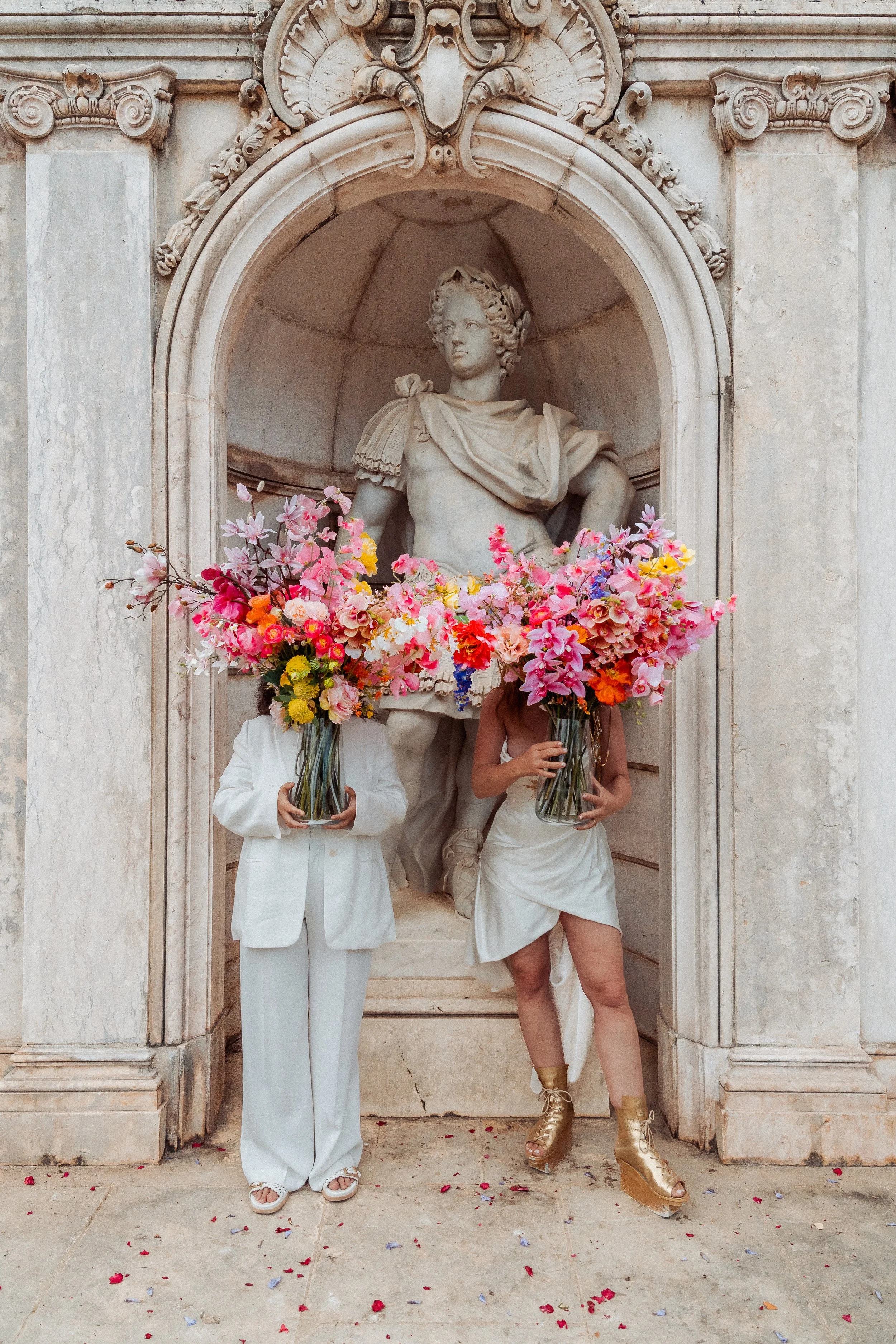 Two individuals dressed in white hold large bouquets of pink, yellow, orange, and purple flowers in front of a marble statue of a historical figure wearing a toga, situated inside an ornate architectural niche.