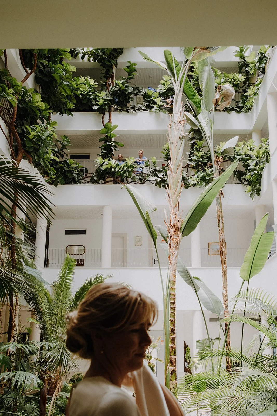 A woman in the foreground with a large indoor garden behind her, featuring tall banana plants and lush green foliage. In the background, two women are standing on an upper balcony of the multi-story building, looking down.