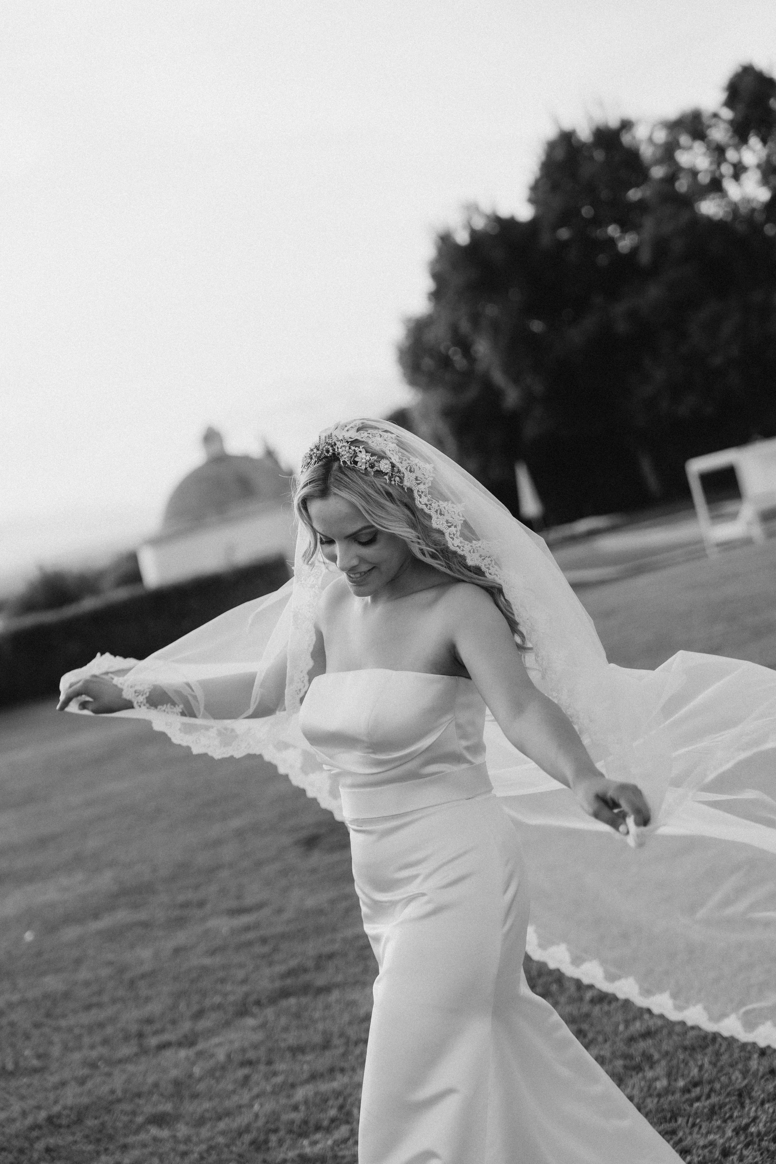 Black and white photo of a bride in a strapless wedding dress, smiling and holding her veil, outdoors on a grassy field with trees and a domed building in the background.