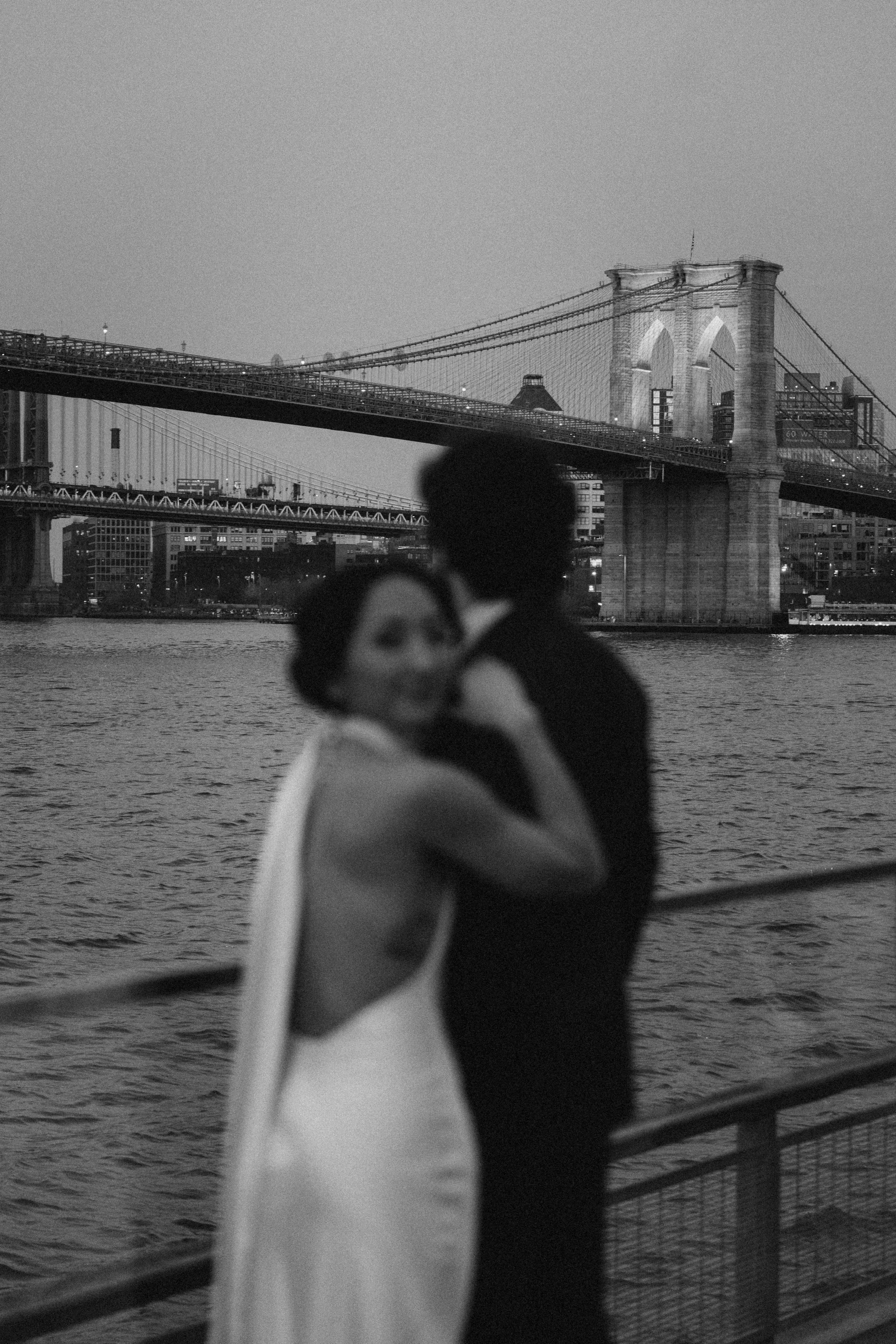 Two people sharing an embrace on a boat with the Brooklyn Bridge and city skyline in the background, in black and white.