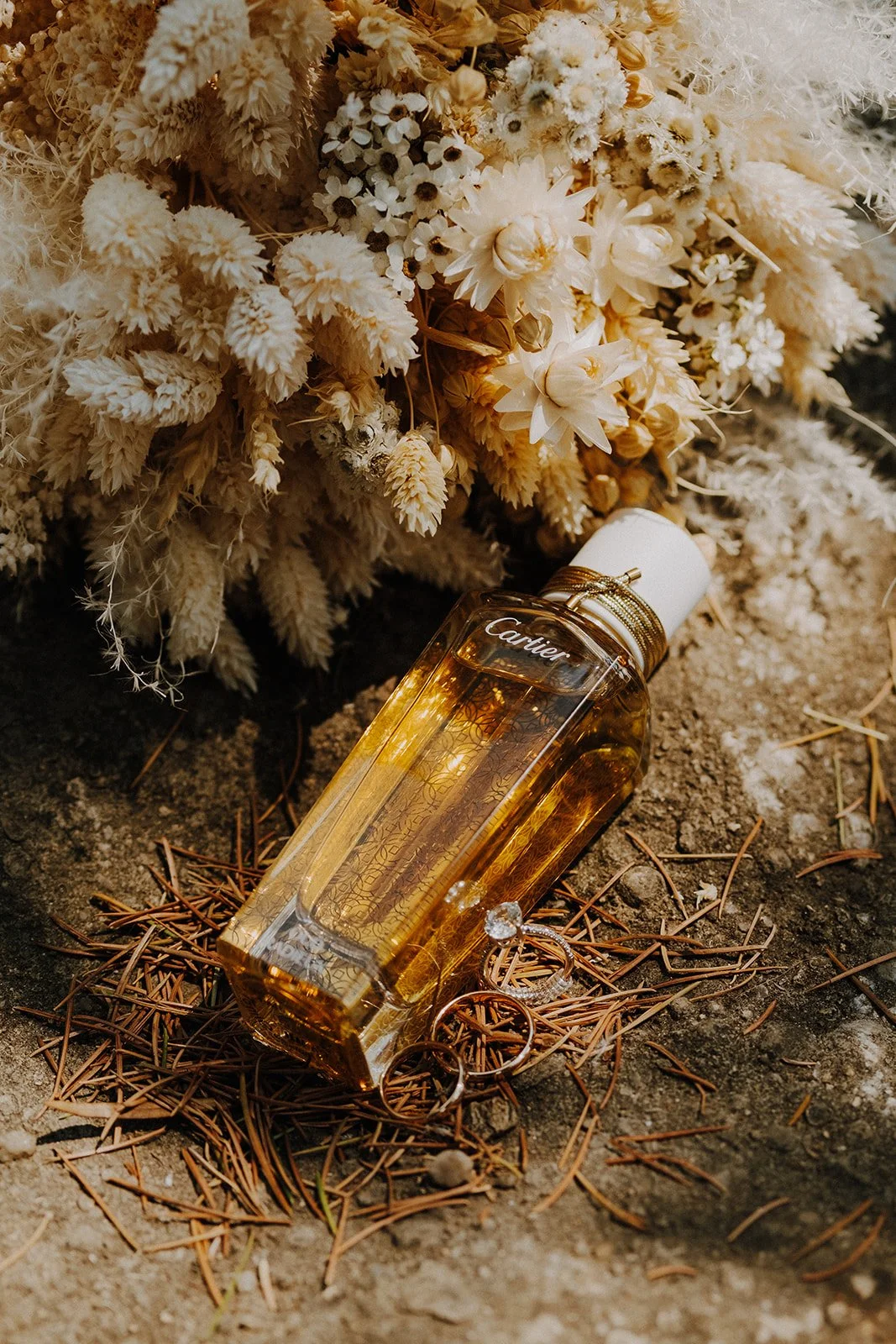 A bouquet of dried white and cream flowers with a bottle of Cartier perfume and a diamond ring on a sandy surface with pine needles.
