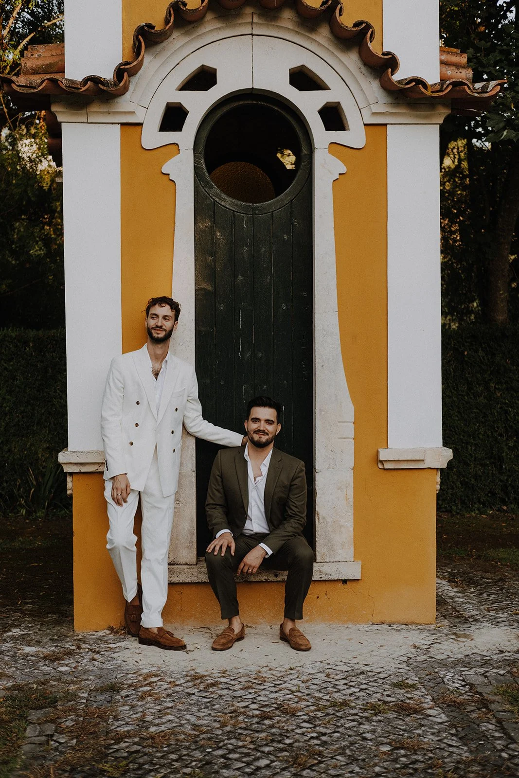 Two men in suits posing in front of a small, ornate yellow and white building with a dark green door and a round window above it.