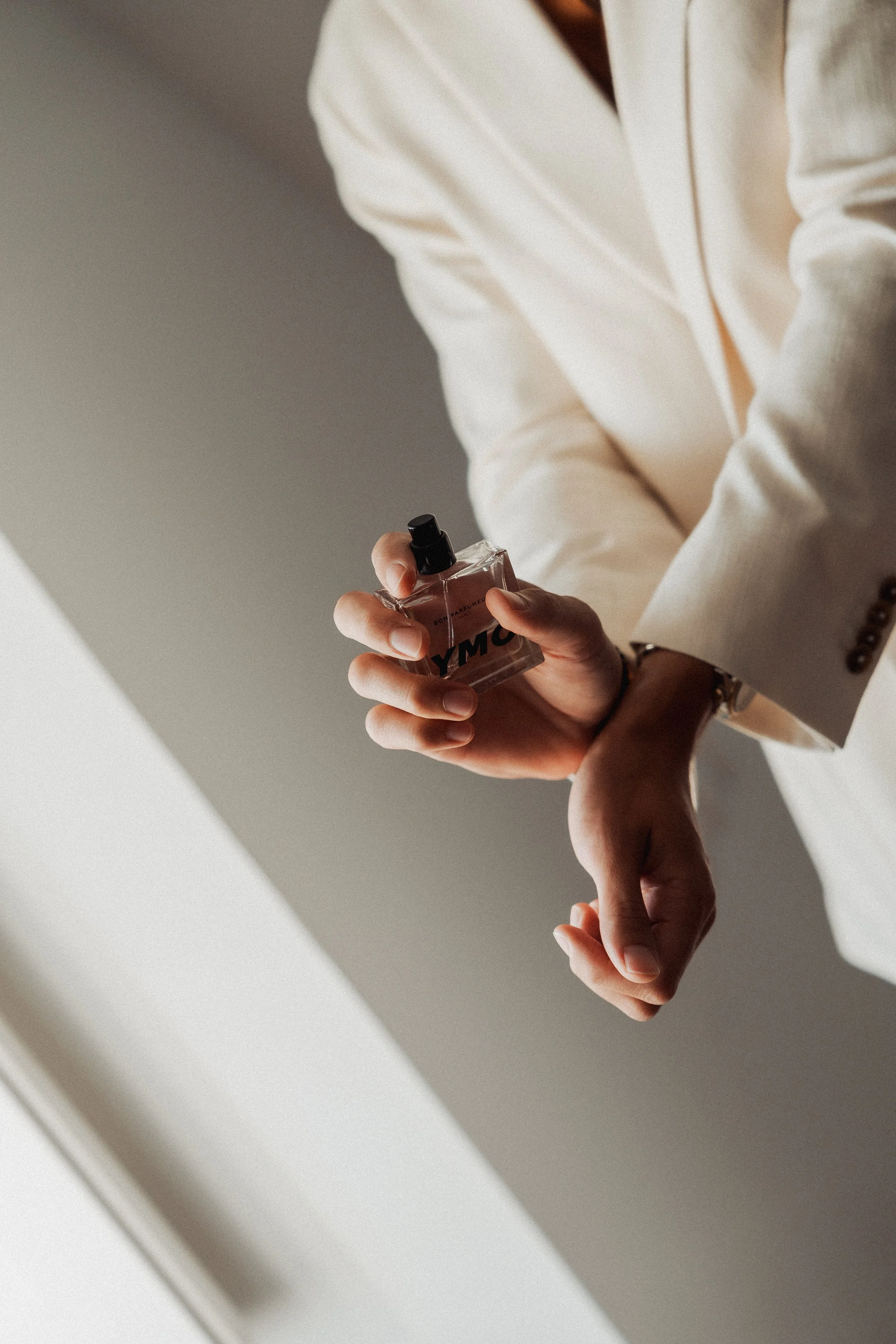 Person dressed in a white blazer holding a perfume bottle labeled 'YMC' in a well-lit indoor setting.
