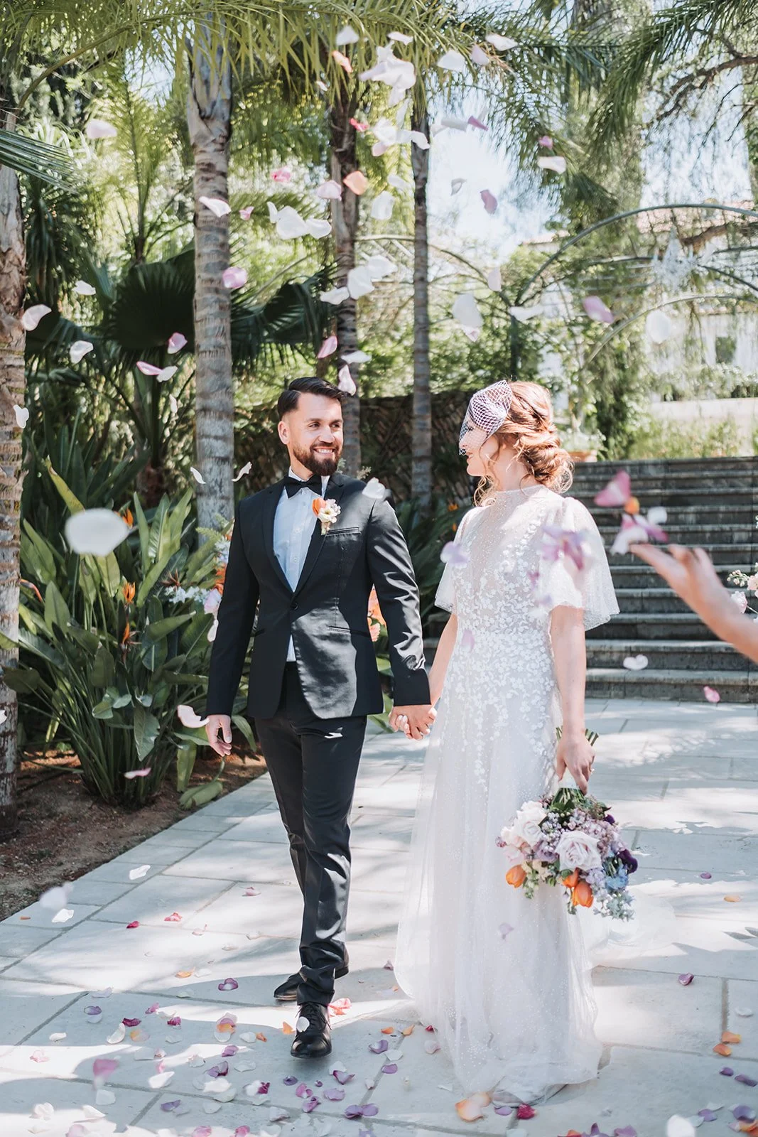 A bride and groom holding hands and walking amid falling flower petals outdoors, surrounded by palm trees and greenery.