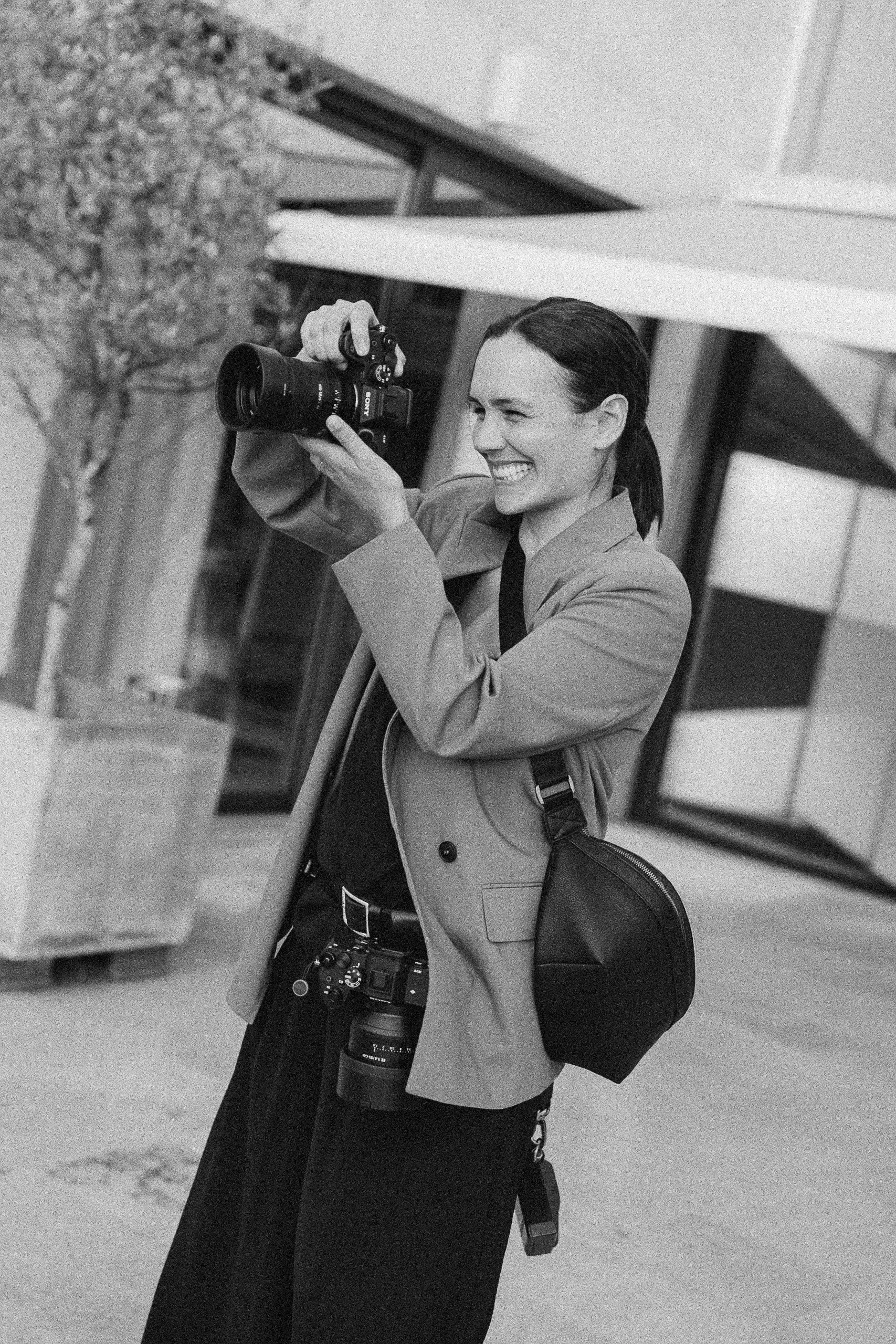 A woman smiling and holding a camera while standing outdoors near a building with a tree in the background.