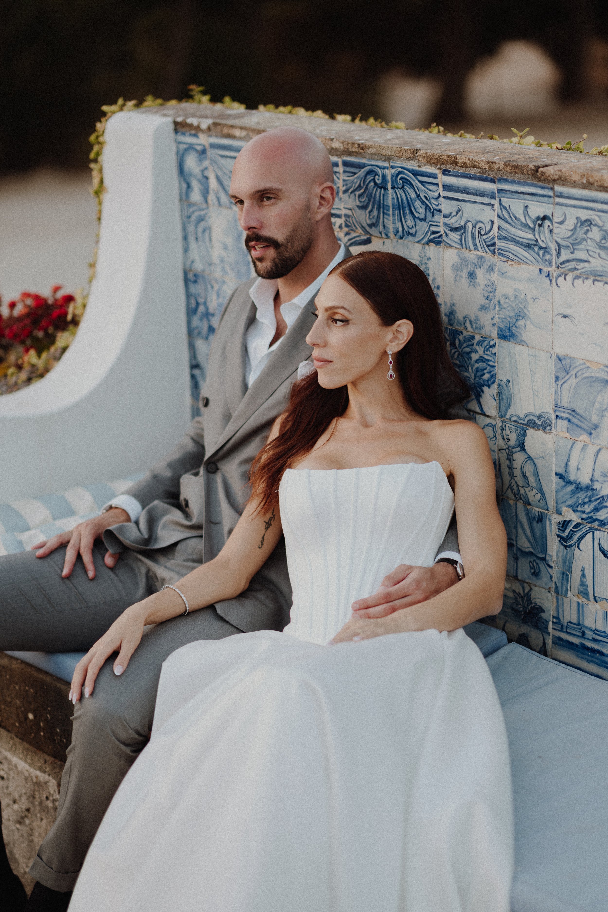 A man and woman in formal attire sitting together outdoors by a decorative blue and white tiled wall during sunset.