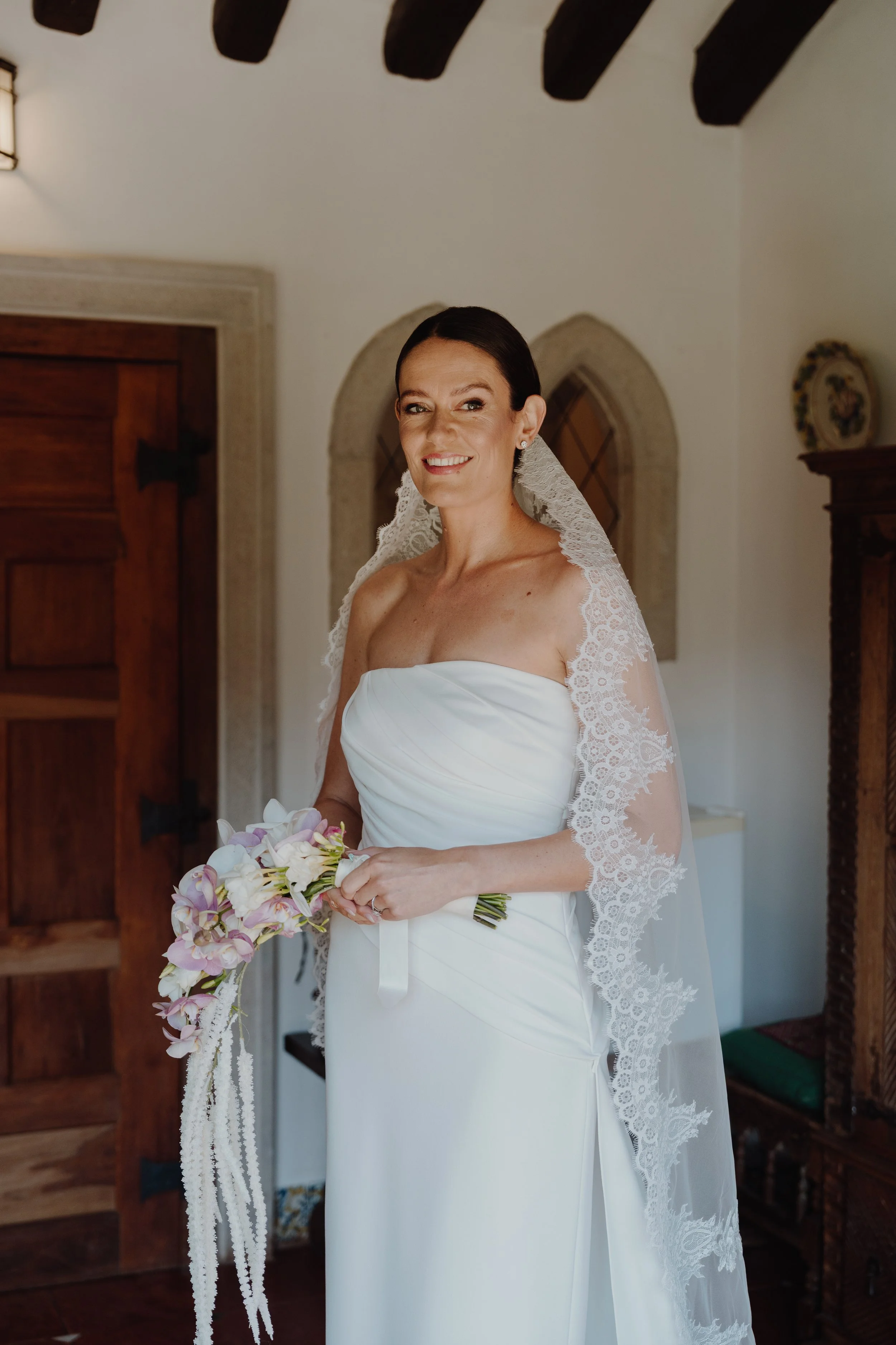 A bride standing indoors, holding a bouquet of pink and white orchids, wearing a strapless white wedding dress with lace veil, smiling at the camera.
