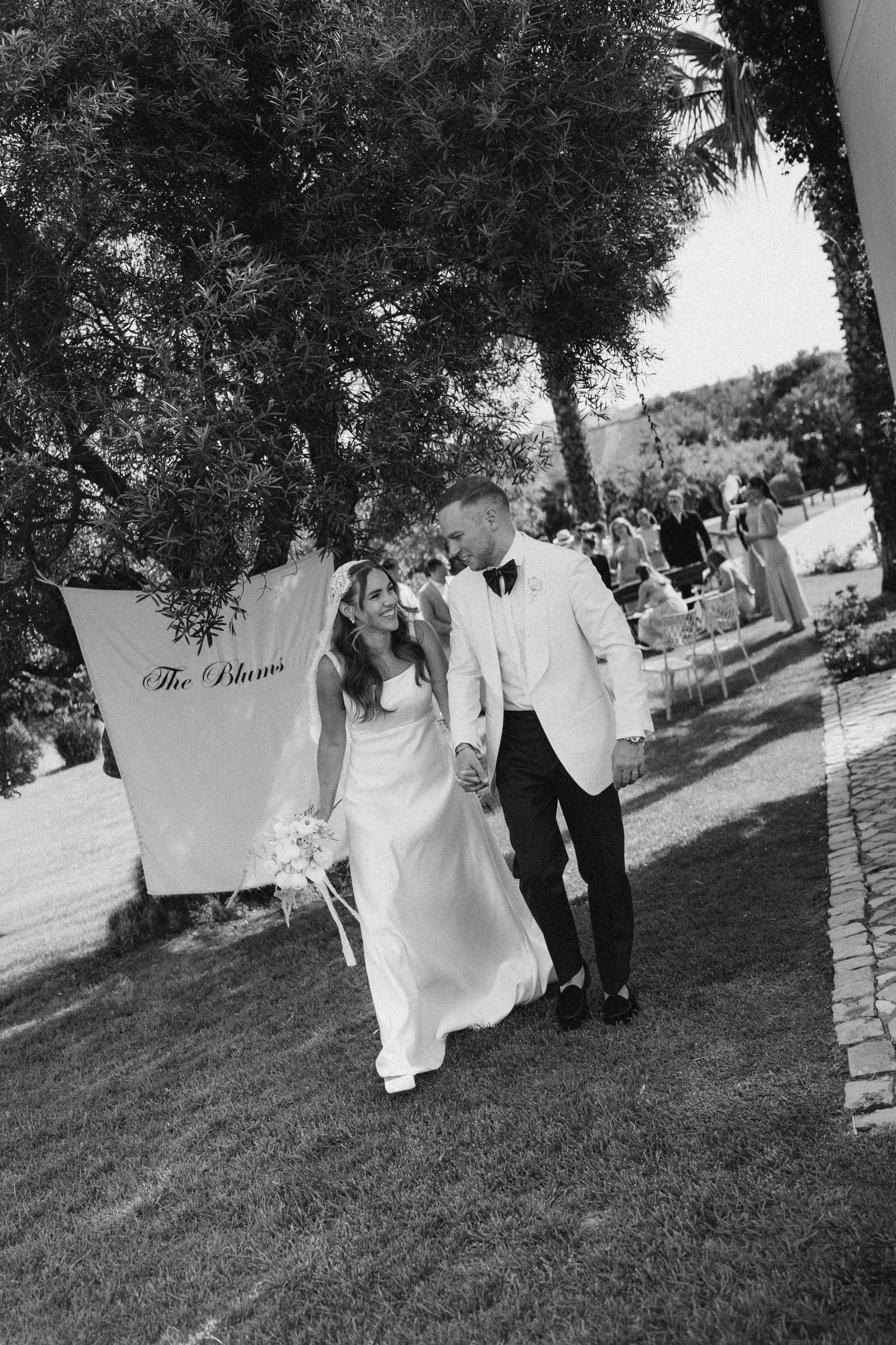 Black and white photo of a bride and groom holding hands and walking outdoors at a wedding reception. The bride is wearing a white gown and a headband with flowers, and the groom is wearing a white tuxedo jacket with a black bow tie. There are guests