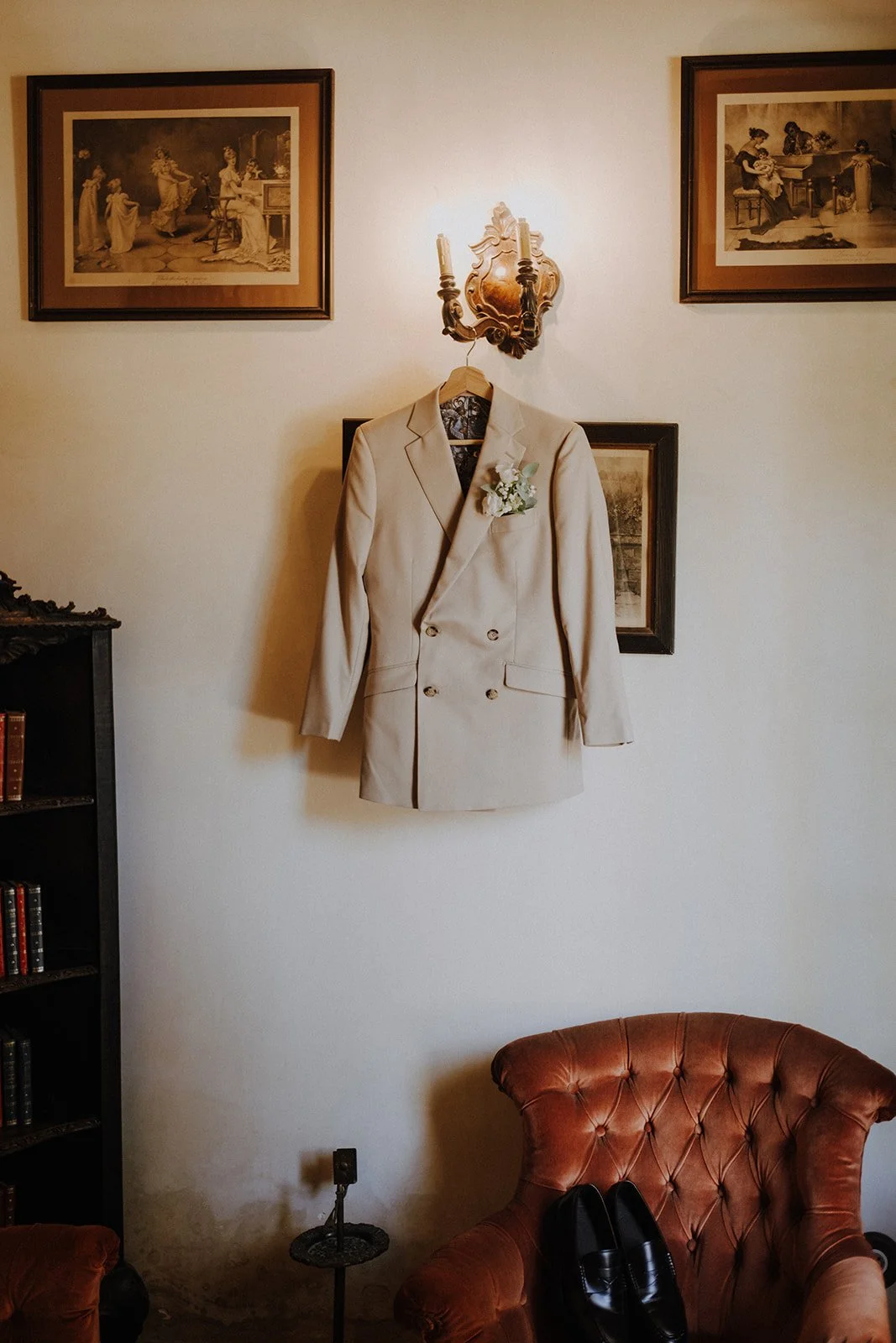 A beige double-breasted suit jacket with a boutonniere hanging on a hanger on a wall with framed vintage pictures, above a brown tufted armchair and a black shoe. A wall sconce light is above the suit.