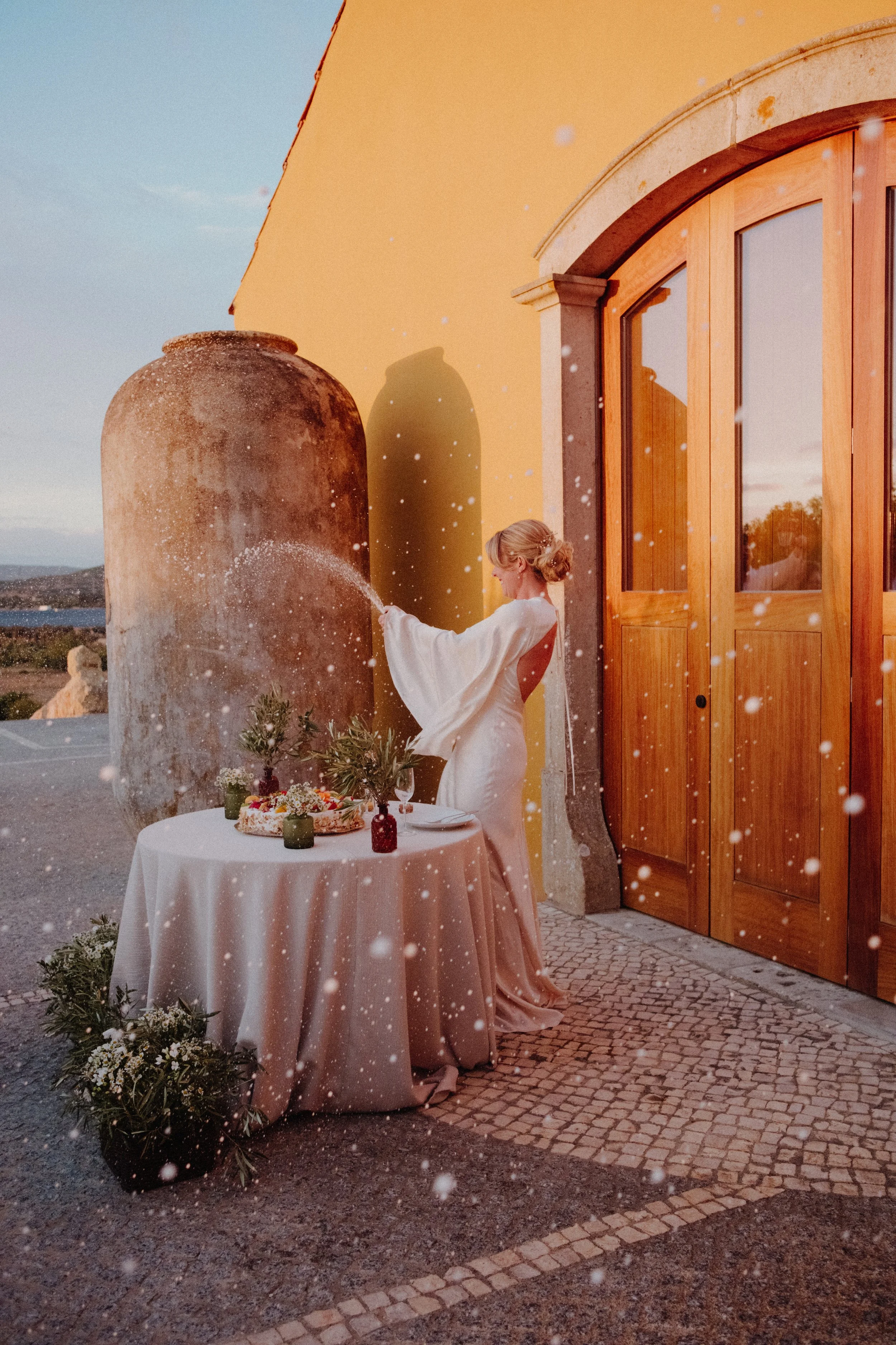 A woman in a wedding dress setting up a table with a cake and flowers outdoors at sunset, with a large yellow building and wooden door in the background, and snow-like particles floating in the air.