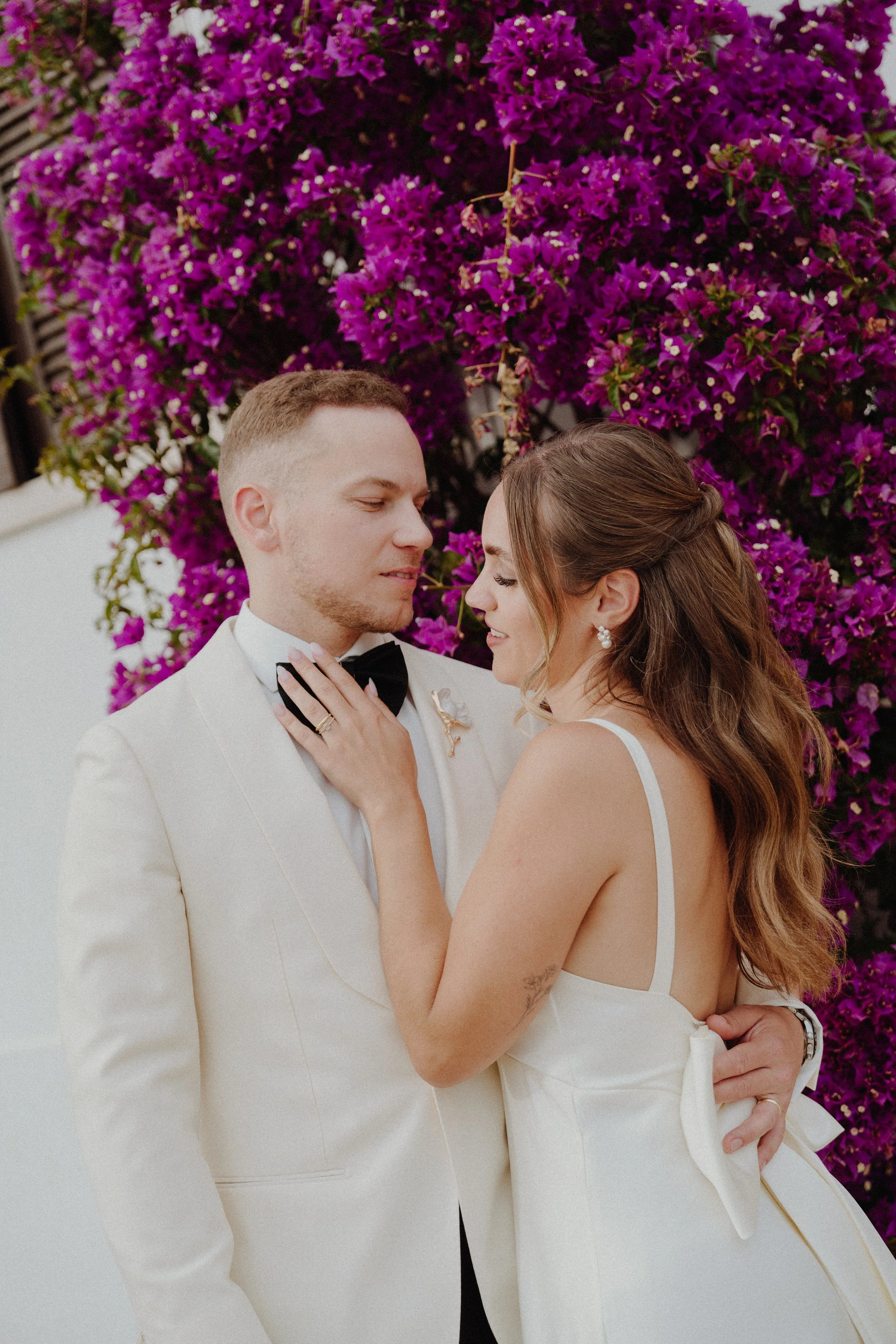 A newlywed couple stands close to each other in front of a vibrant purple bougainvillea flower background. The groom is dressed in a white tuxedo with a black bow tie, and the bride is wearing a white wedding gown with spaghetti straps and pearl earrings. They are gazing into each other's eyes, with the bride's hand gently placed on the groom's chest, showing her wedding ring.