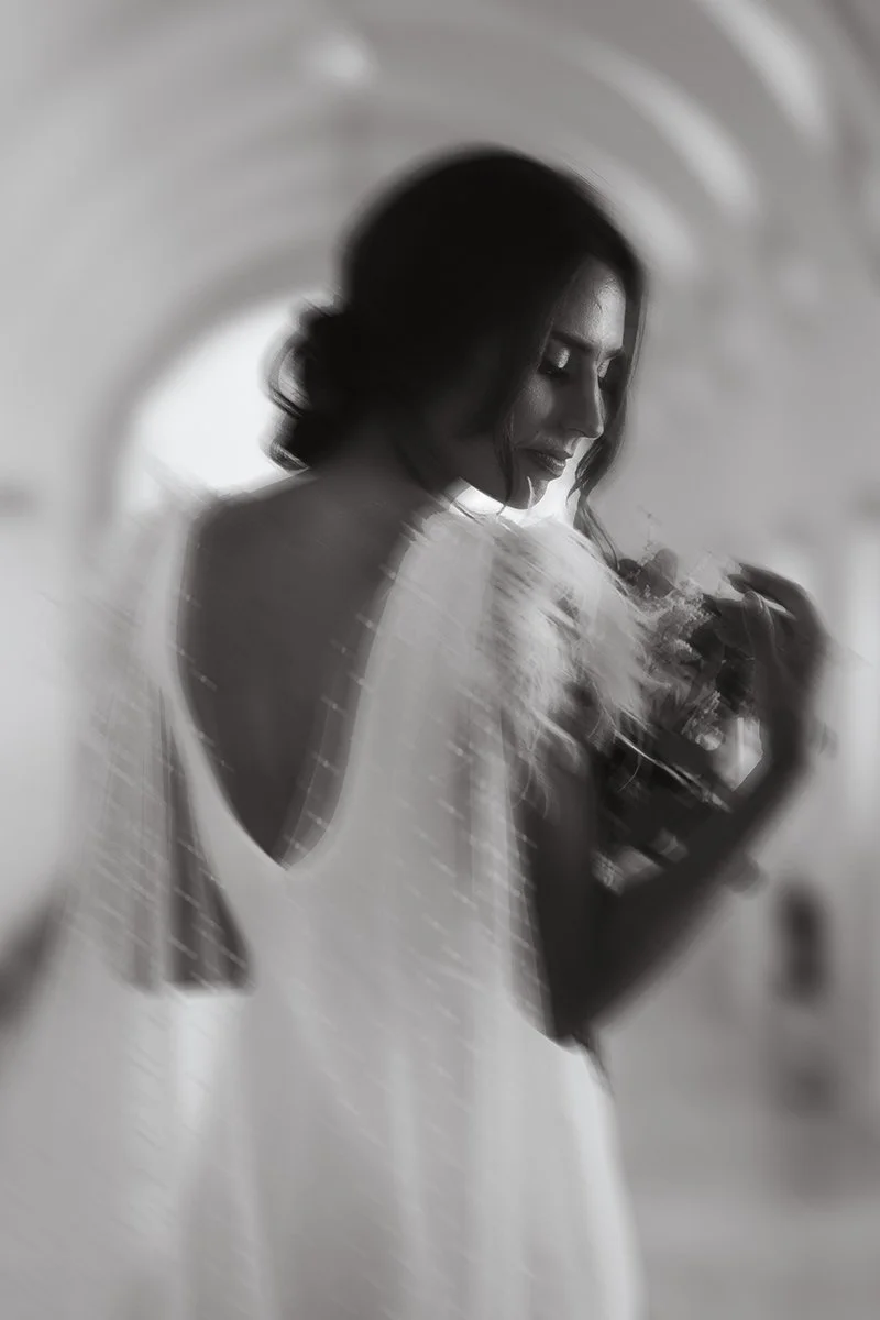 Black and white photo of a woman looking at her phone, with blurred city lights reflected over her. The woman has dark, wavy hair and is wearing a white dress with an open back.