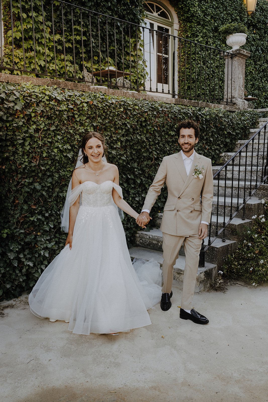 A bride and groom holding hands and smiling outdoors on their wedding day, with greenery and stairs in the background.