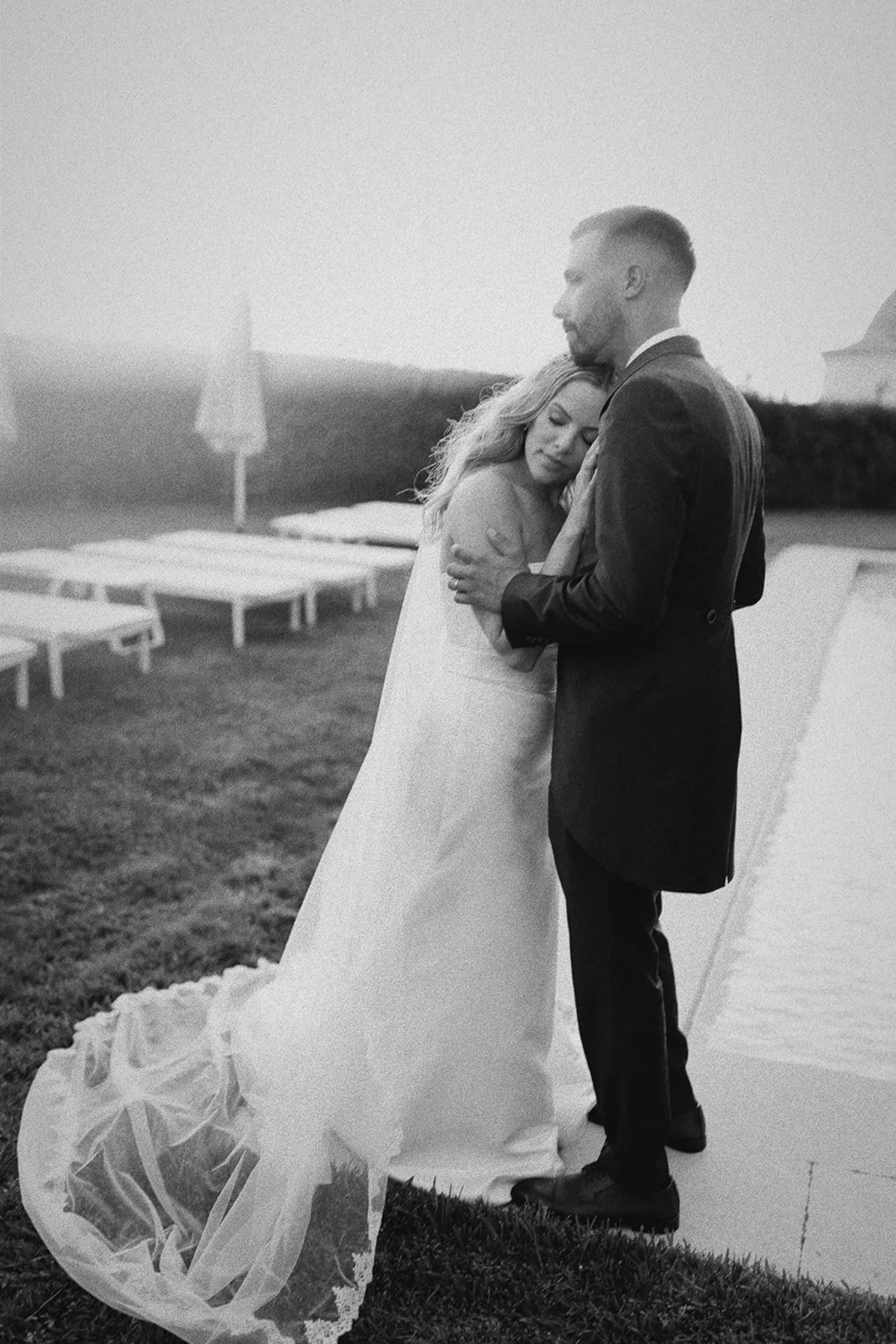 A bride and groom embracing outdoors during sunset on their wedding day, with empty lounge chairs and umbrellas in the background.