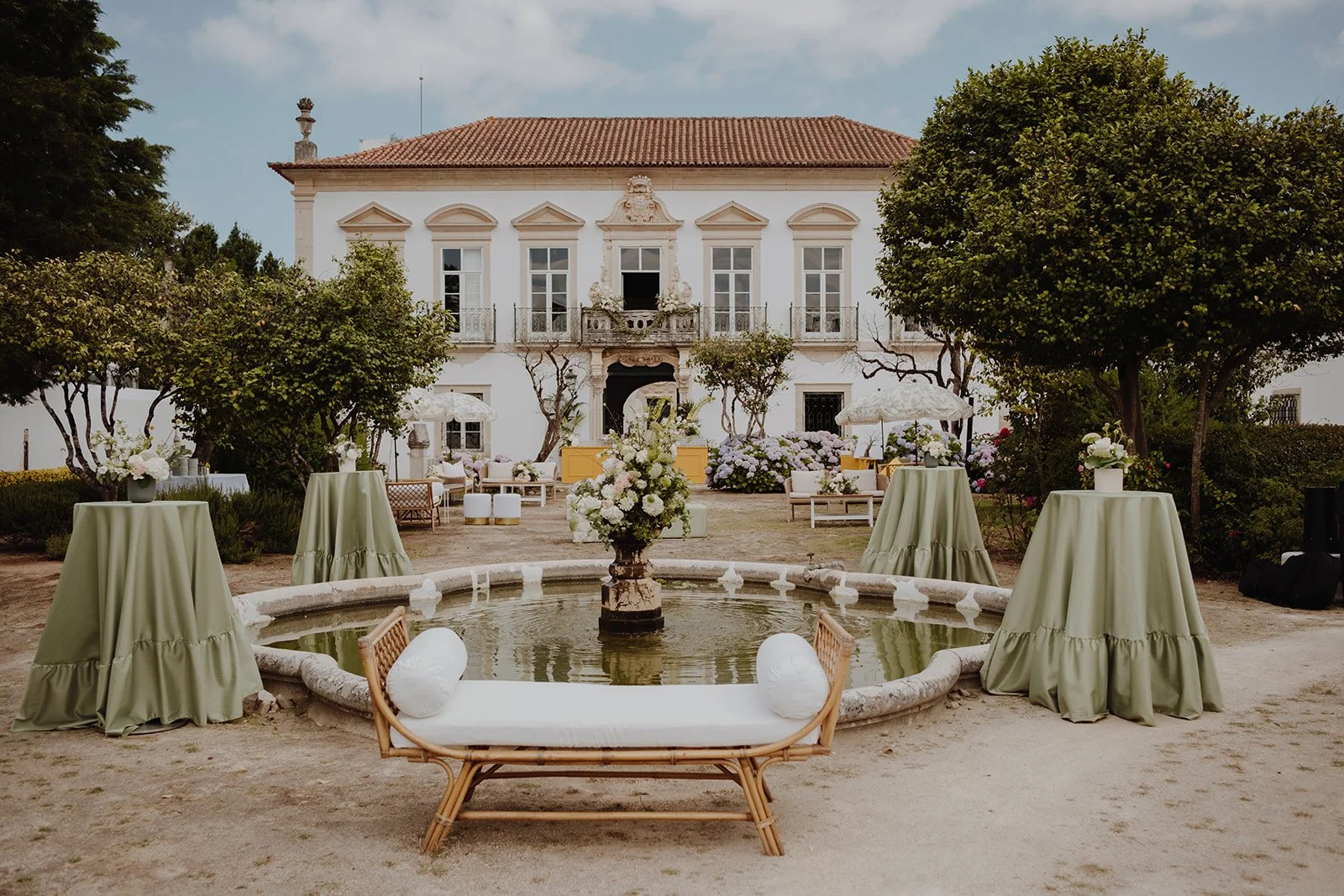 Elegant outdoor courtyard with a central fountain decorated with flowers, surrounded by tables with green tablecloths, umbrellas, and seating, in front of a large white historic building with multiple windows and a red-tiled roof.