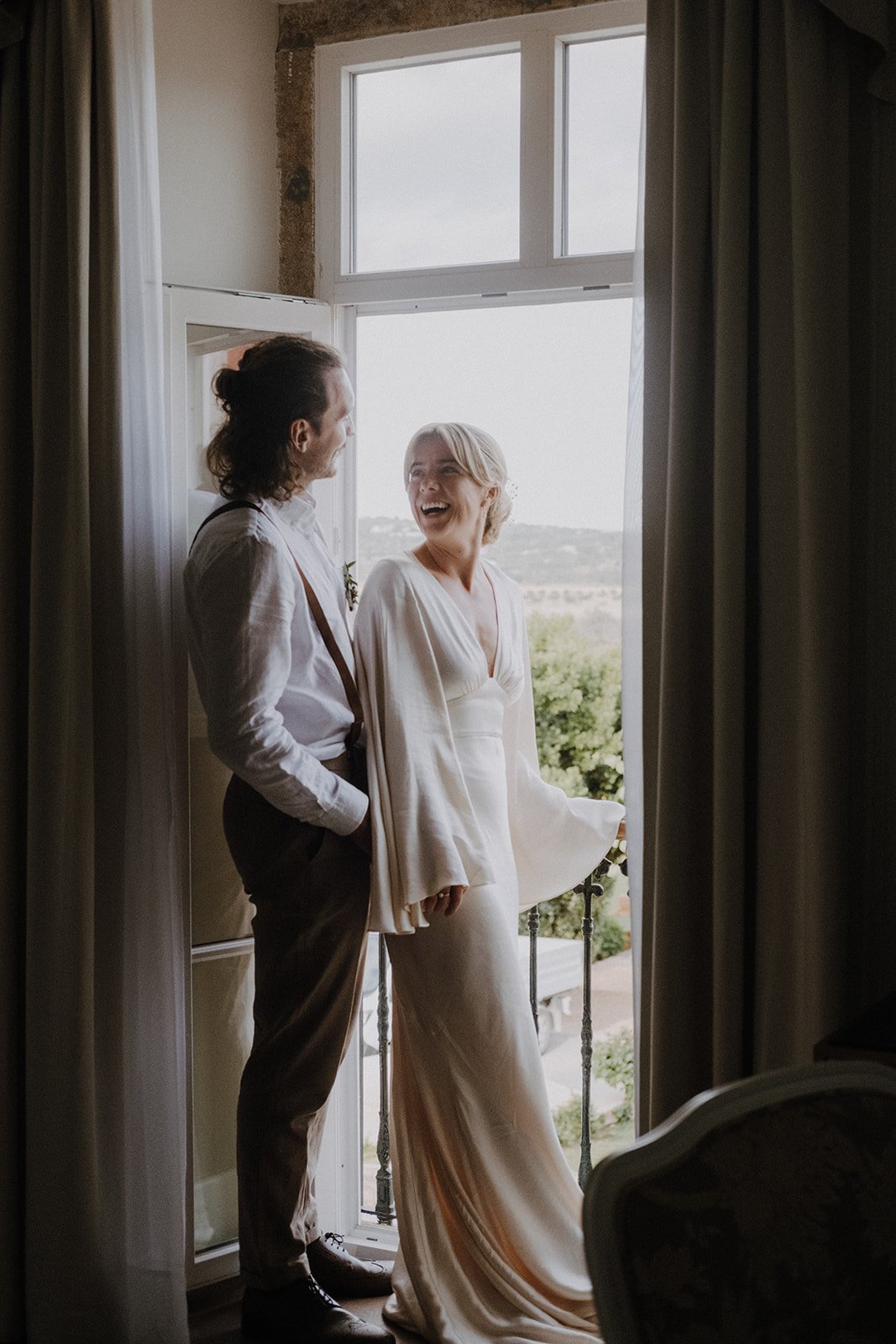 A couple standing by an open window, smiling and looking at each other, with scenic outdoor view in the background. The woman is in a white dress, and the man is in a white shirt with suspenders.