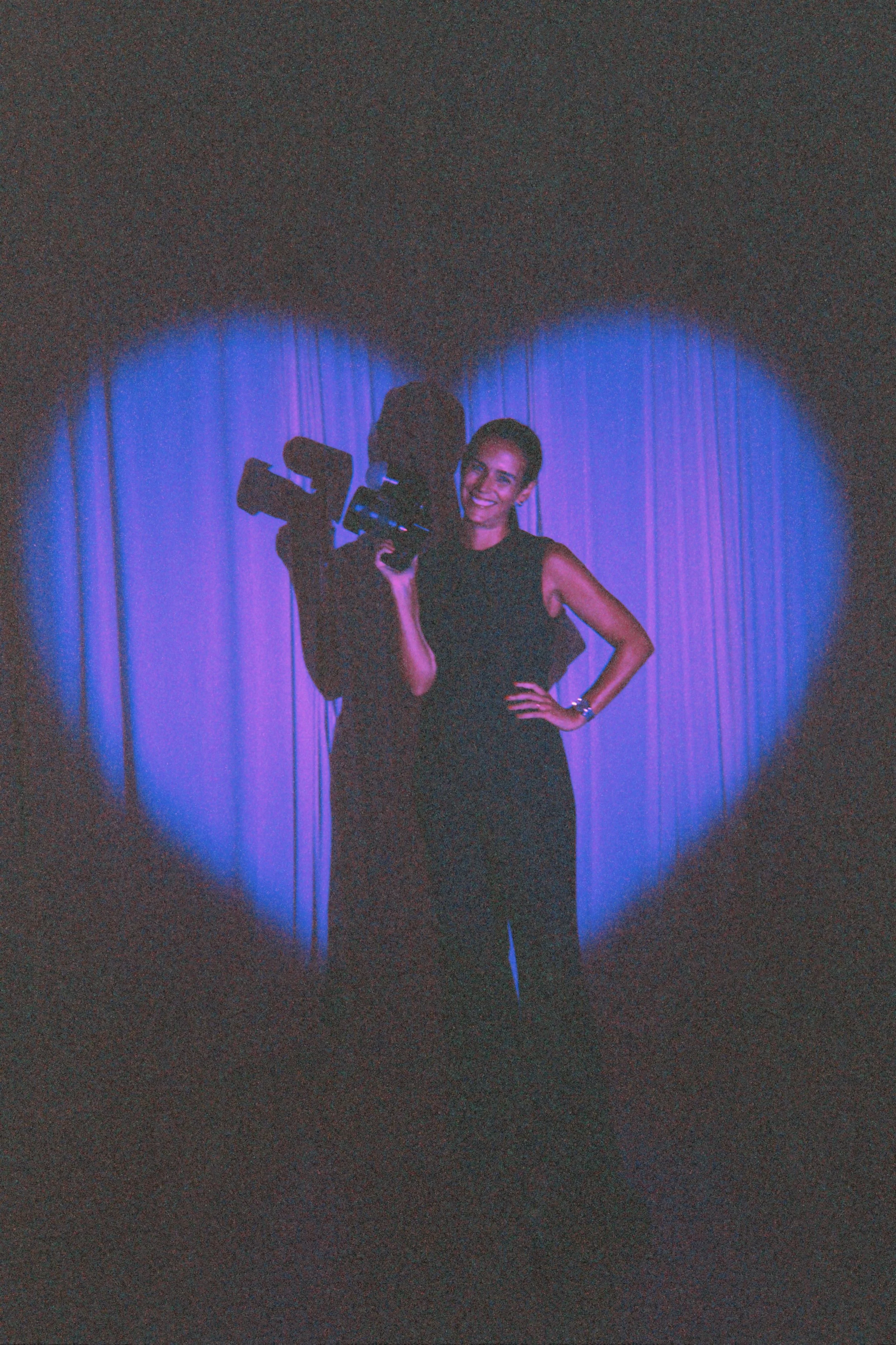 A person with short dark hair wearing a black sleeveless top, smiling, and holding a video camera, standing in front of a purple curtain, illuminated by a spotlight.