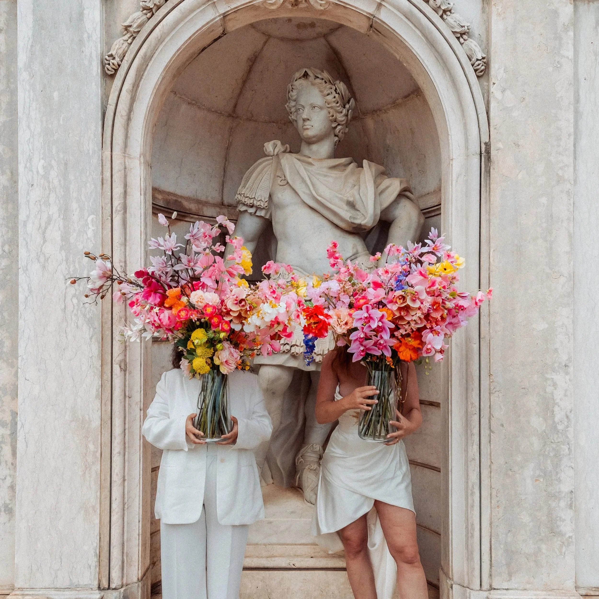 Two people holding large flower bouquets standing in front of a beige stone wall with a marble arch behind a statue of a woman in classical attire in an alcove.