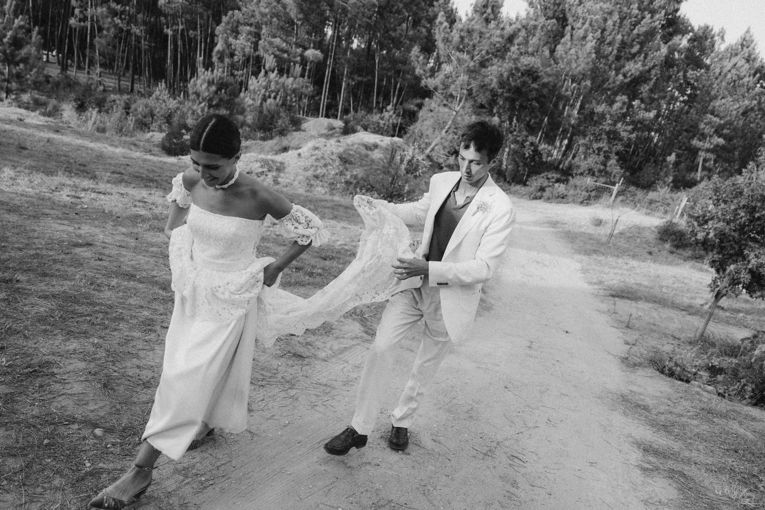 A black and white photo of a couple walking along a dirt path in a natural outdoor setting. The woman is wearing an off-shoulder lace wedding dress, and the man is dressed in a light-colored suit. They are holding hands, and the woman is lifting her 