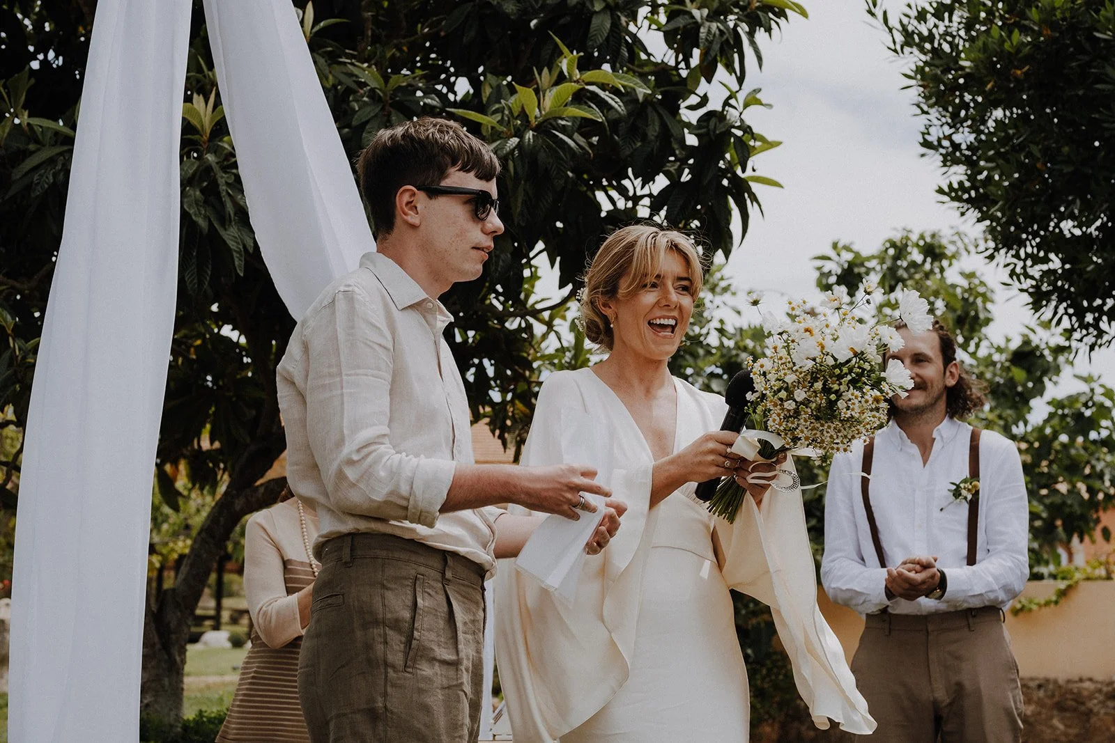 A woman in a white dress holding a bouquet of flowers and a microphone, smiling at a wedding ceremony outdoors, with two men in dress shirts and suspenders nearby, under a white fabric arch surrounded by trees.