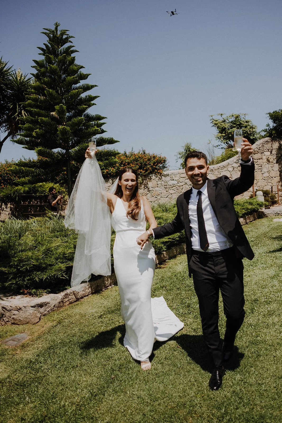 A newlywed couple celebrating outdoors on a sunny day, with the bride in a white wedding dress and veil, and the groom in a black suit and tie. They are holding glasses and smiling, with a drone flying above them and a person taking photos in the bac