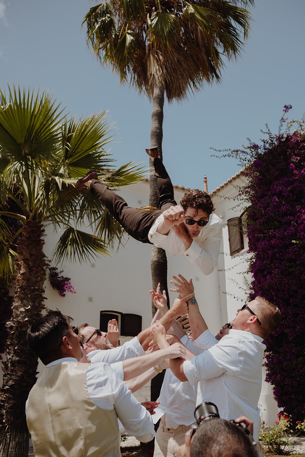 A man wearing a white shirt and sunglasses is being tossed into the air by a group of people outdoors, with palm trees and purple flowers surrounding them.