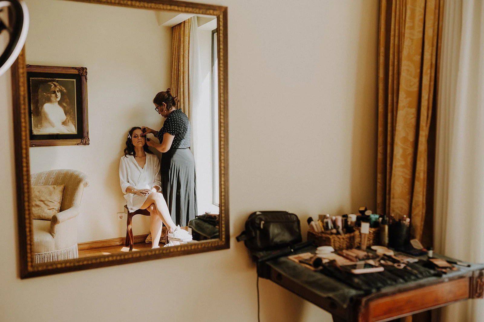 A woman in a white robe sitting on a chair, getting her makeup done by a makeup artist in a room with a window and yellow curtains, reflected in a mirror, with makeup supplies on a nearby table.