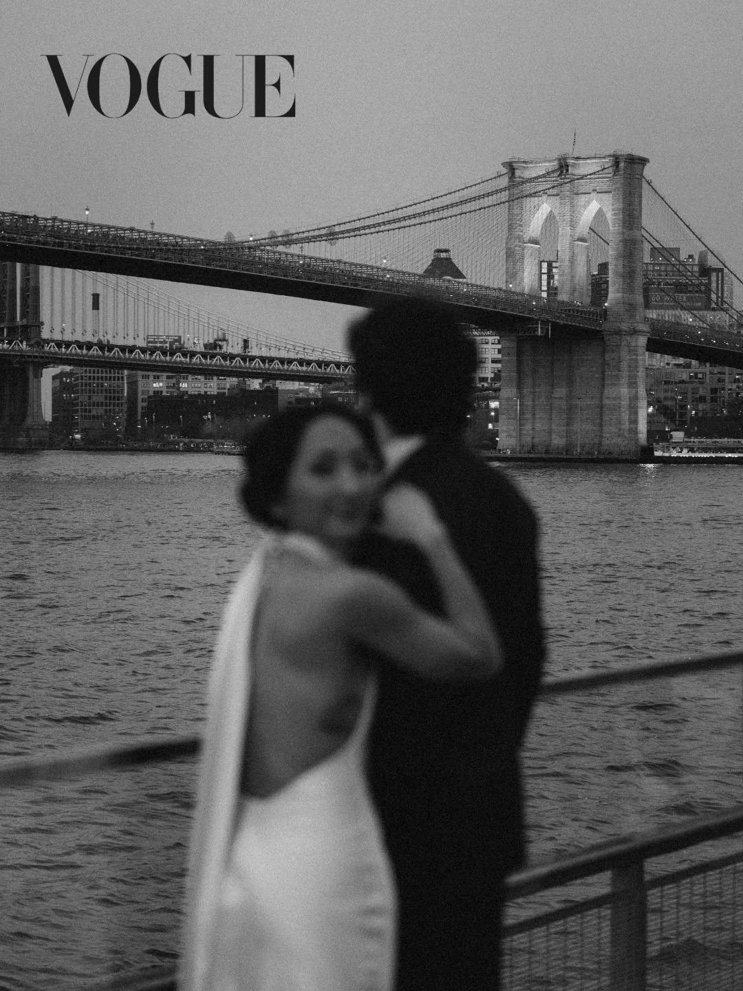 Black and white photo of a couple embracing by the water with the Brooklyn Bridge in the background.