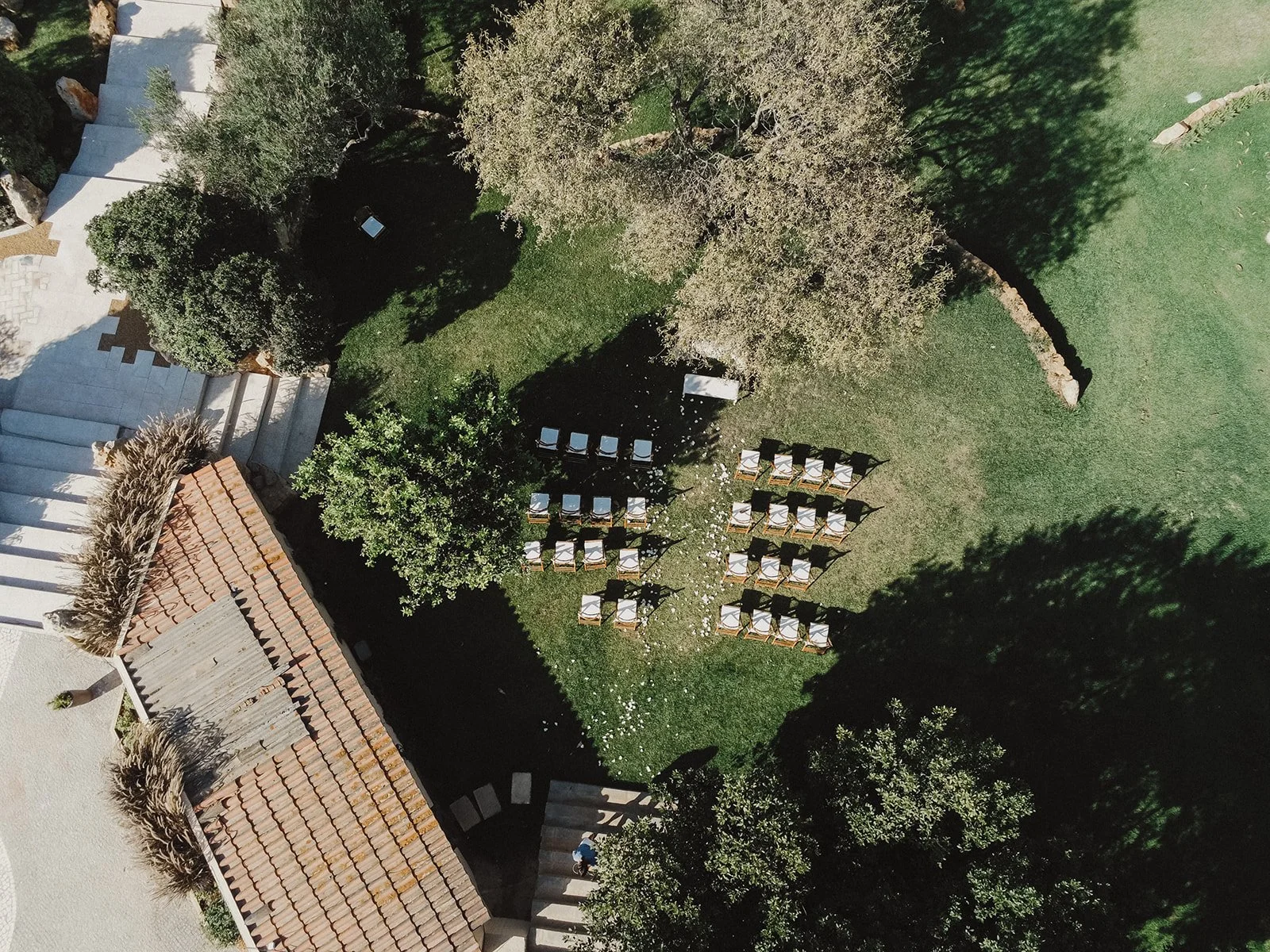 Aerial view with drone an outdoor wedding setup with multiple rows of chairs on a grassy area, surrounded by trees and a building with a tiled roof.