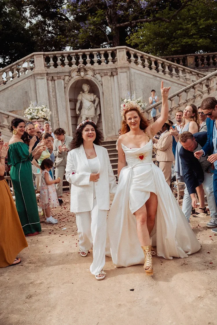 A gay wedding celebration outdoors with two women in the center, one in a white groom's suit and the other in a wedding dress, surrounded by friends applauding and smiling. The scene features a grand staircase with classical statue and floral decorat