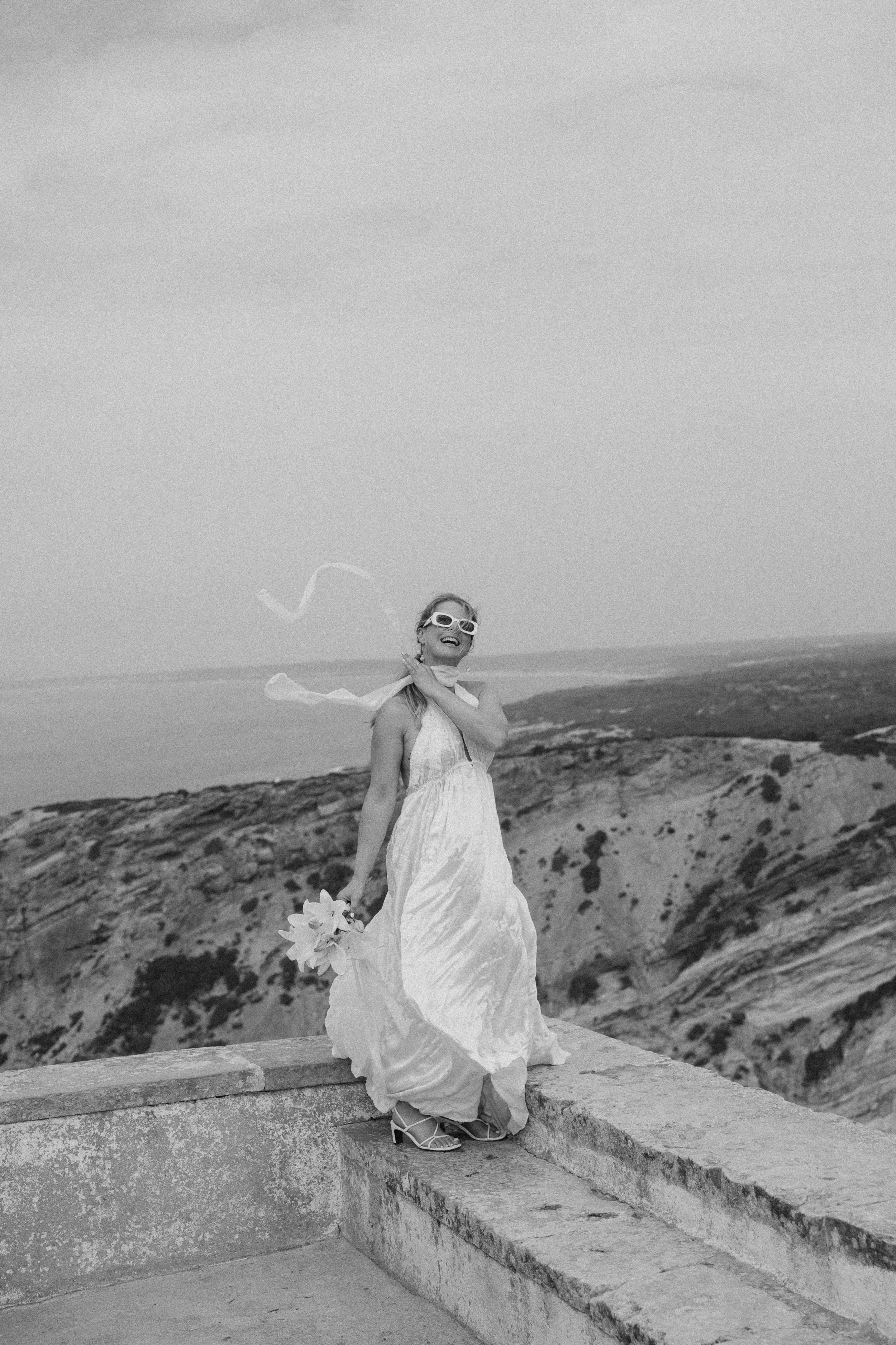 A woman in a wedding dress standing on stone steps outdoors, holding a bouquet of flowers, with natural landscape background and wearing sunglasses.