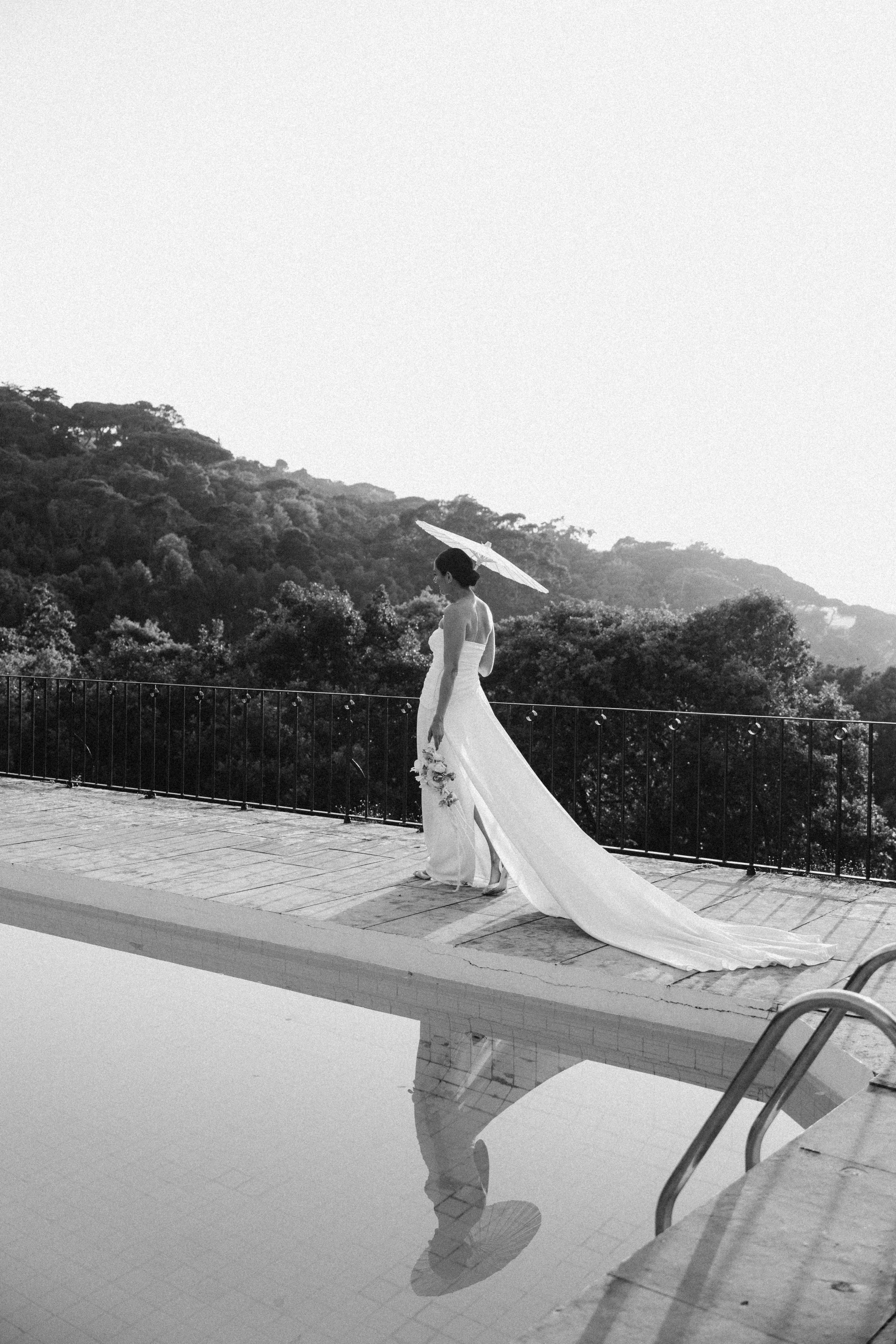 A woman in a wedding dress standing by a swimming pool, holding a bouquet and an umbrella, with a scenic hilly landscape in the background, reflected in the pool water, in black and white. Quinta São Thiago.