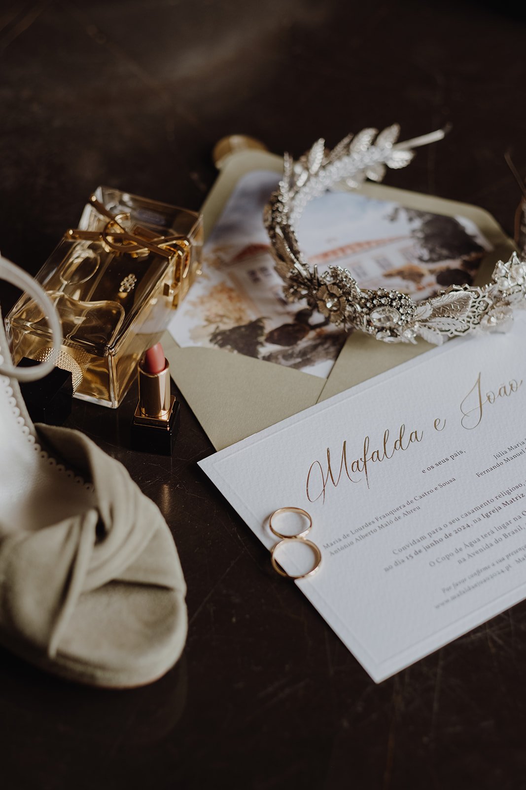 Wedding invitation with two gold wedding bands, a tiara, lipstick, perfume, a beige headband, and wedding photos on a dark wooden surface.