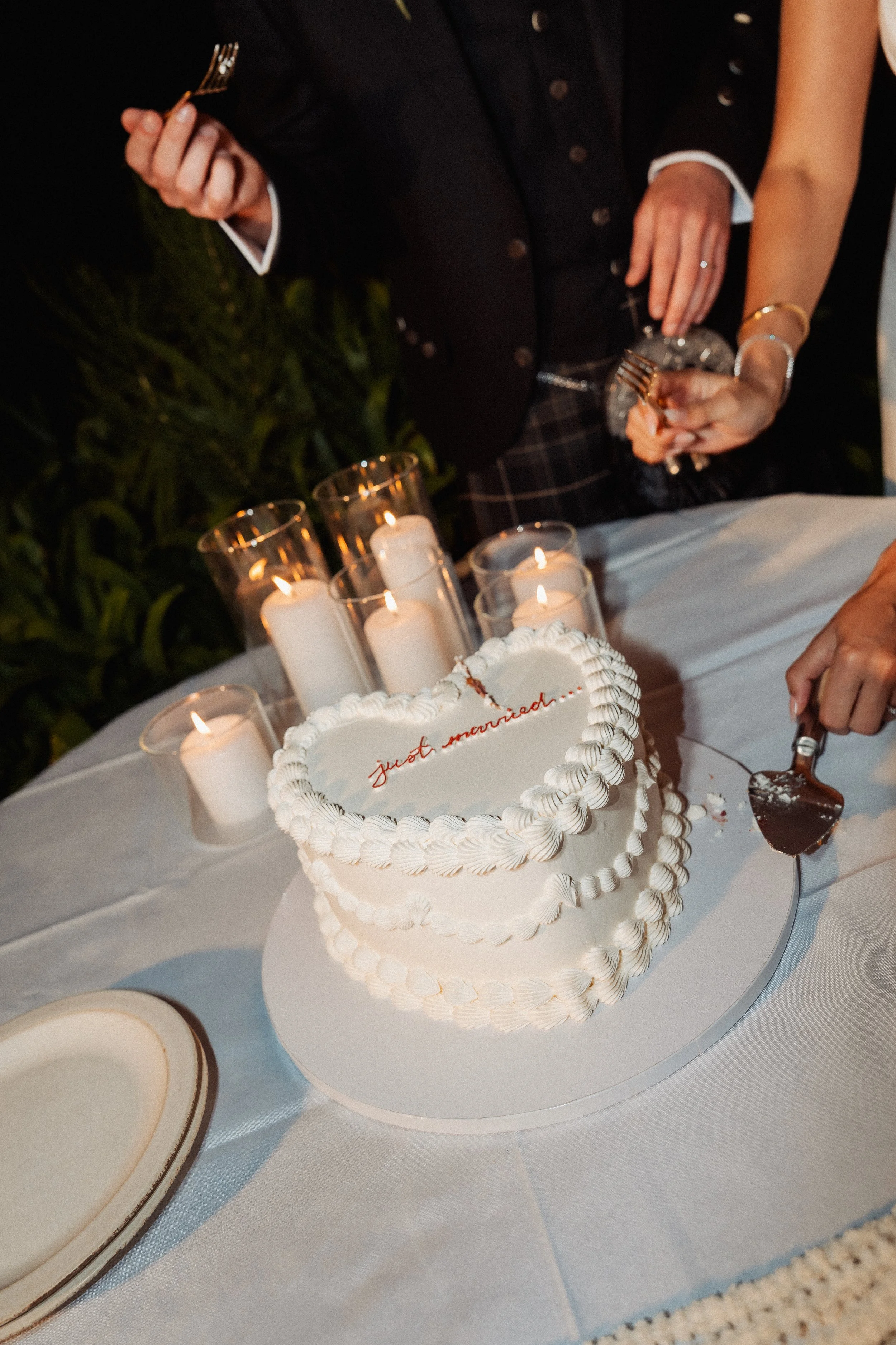 Wedding cake with the message "just married" written on it, surrounded by candles, with two people cutting the cake at a celebration.