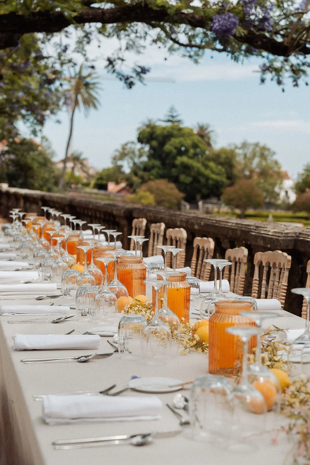 Long banquet table set outdoors with white tablecloth, orange and clear glass jars, white plates, silver cutlery, and white napkins, under a tree with purple flowers in a garden setting. Vandeli Gardens. 