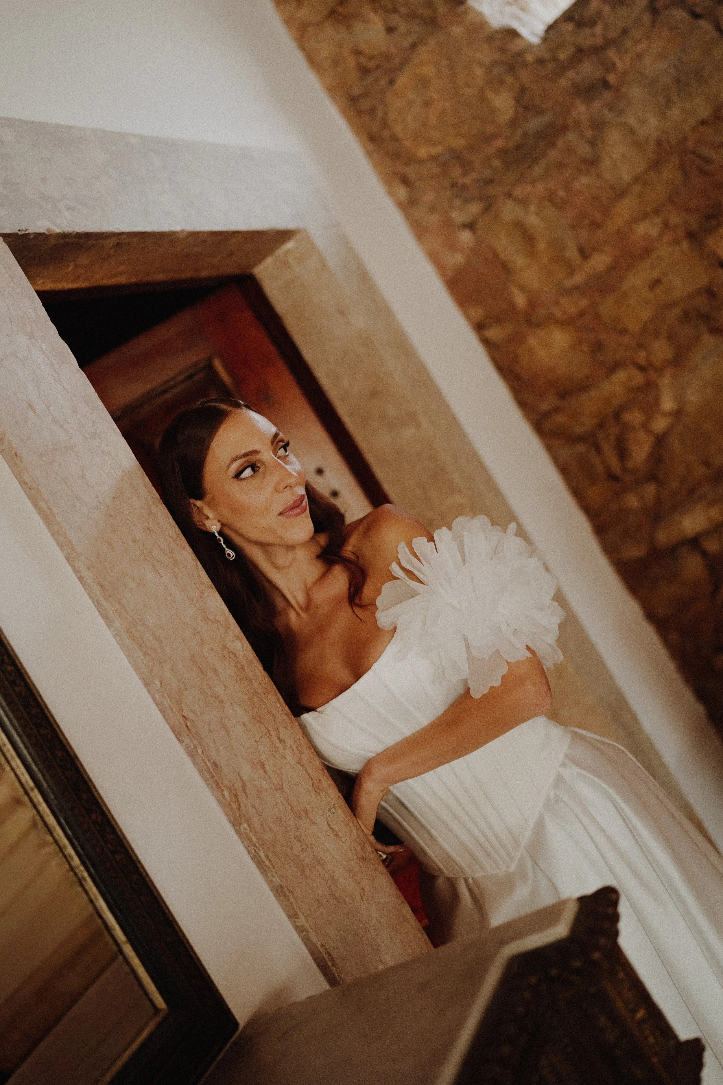 A woman in a white dress with floral details holds a bouquet of white flowers, standing in front of a stone wall and doorway, with a mirror reflecting part of her face.