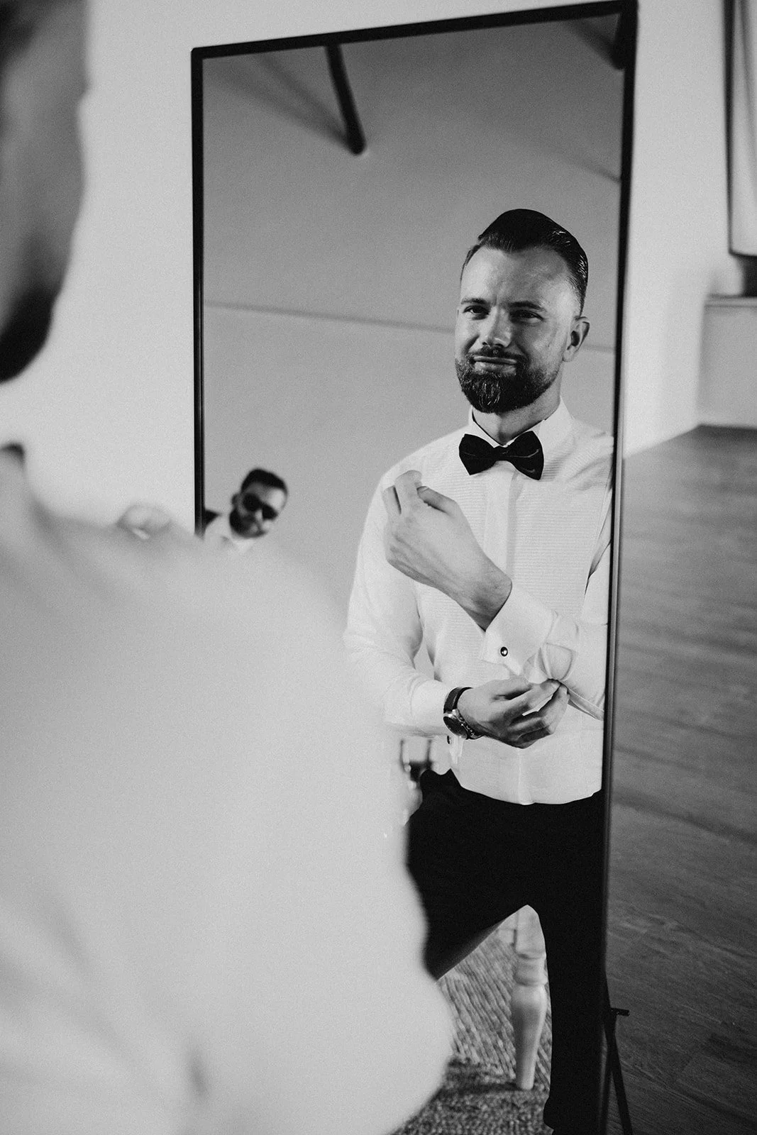 A man dressed in formal attire with a bow tie, seen through a mirror, adjusting his cuff while another man in the background wearing sunglasses looks on.