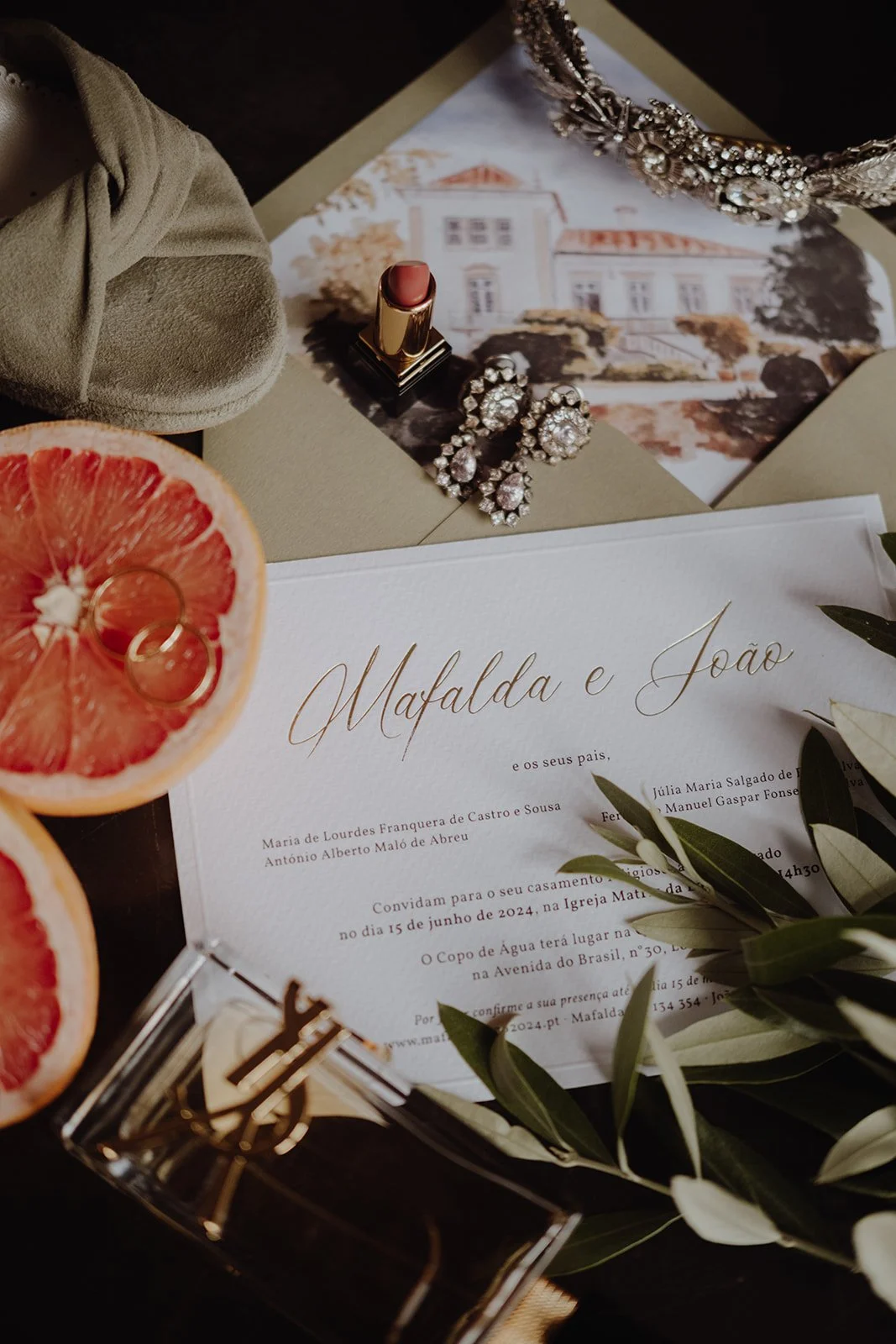 Wedding invitation with grapefruit slices, jewelry including rings, earrings, and a brooch, lipstick, and greenery.