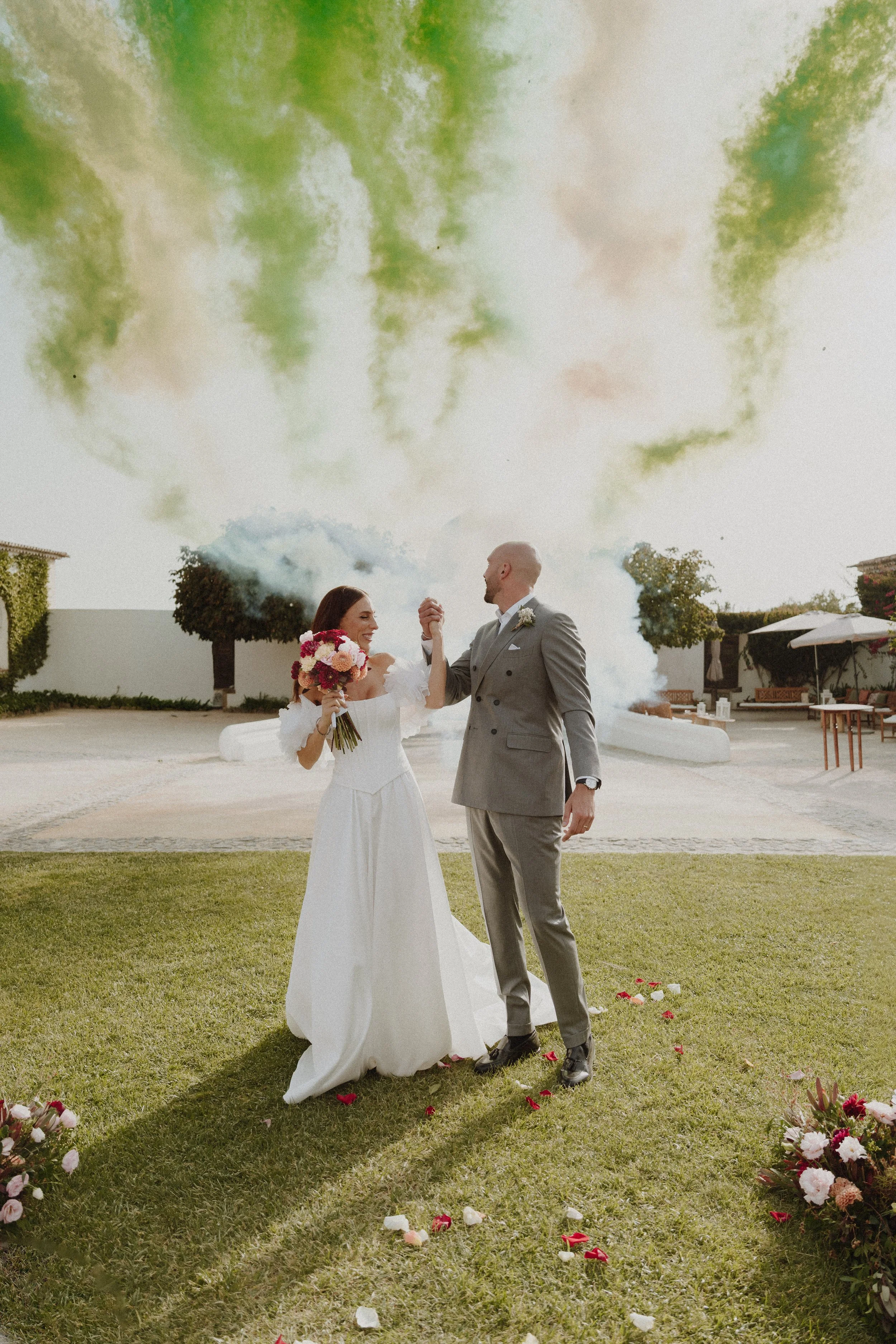 A bride and groom stand on a grassy area at their wedding ceremony, holding hands, with confetti and flower petals on the ground, and smoke bombs in the background creating colorful clouds in the sky.