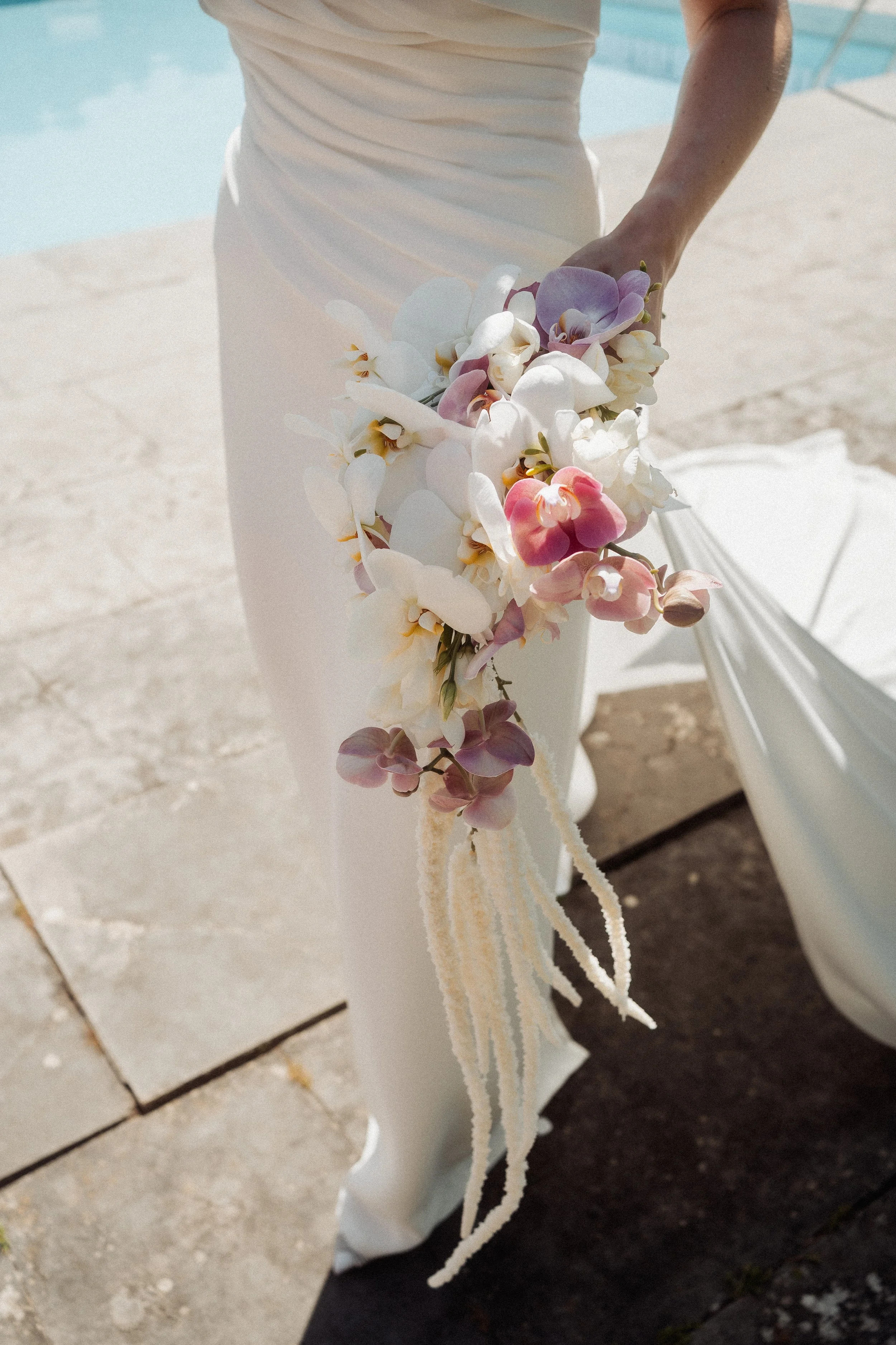 Bride in a white dress holding a cascading bouquet of white, pink, purple, and peach orchids near a pool on a sunny day.