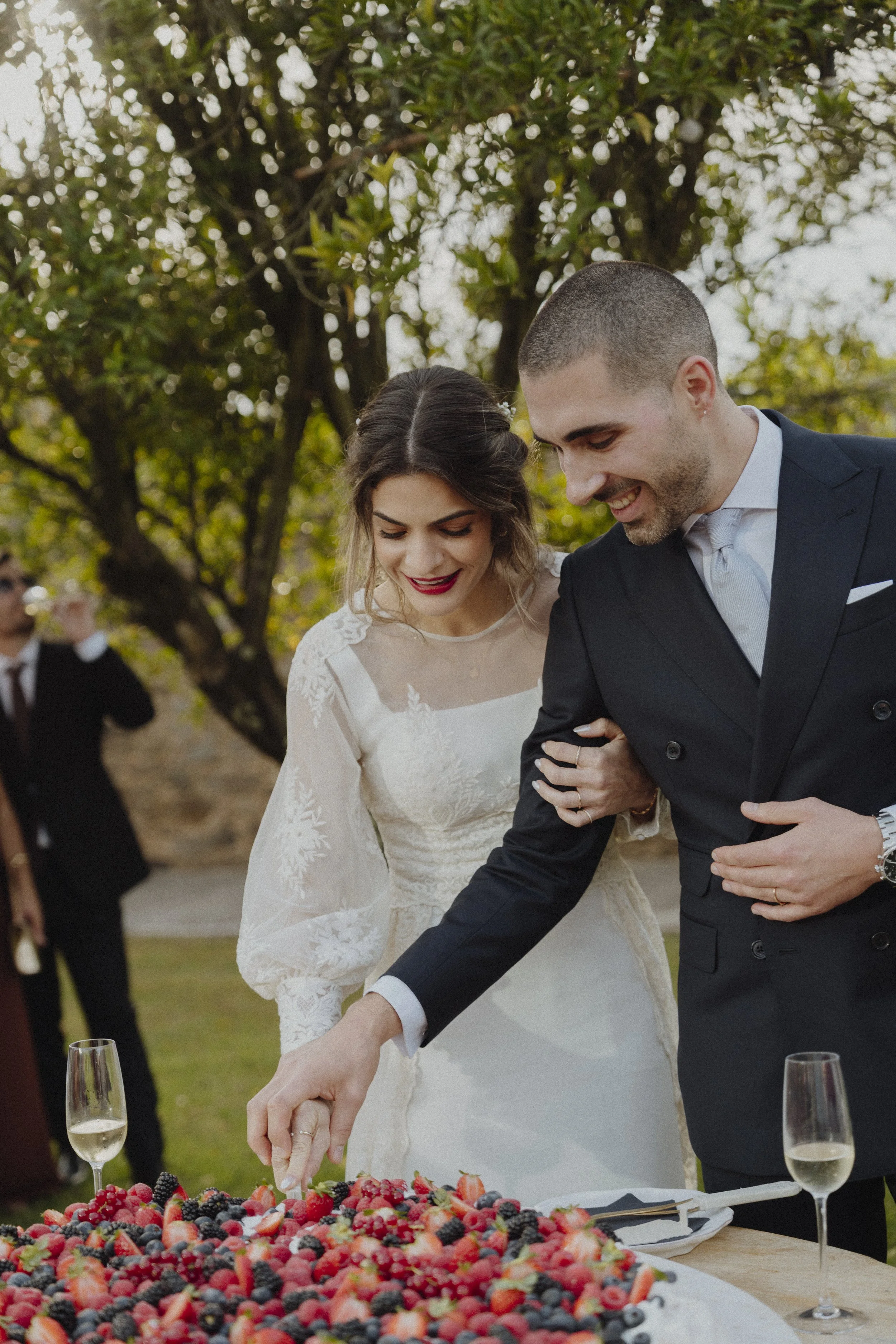 A bride and groom at their wedding reception cutting a large tray of assorted berries with wine glasses nearby. The bride is wearing a white lace wedding dress, and the groom is dressed in a black suit with a white shirt and light tie, outdoors with 