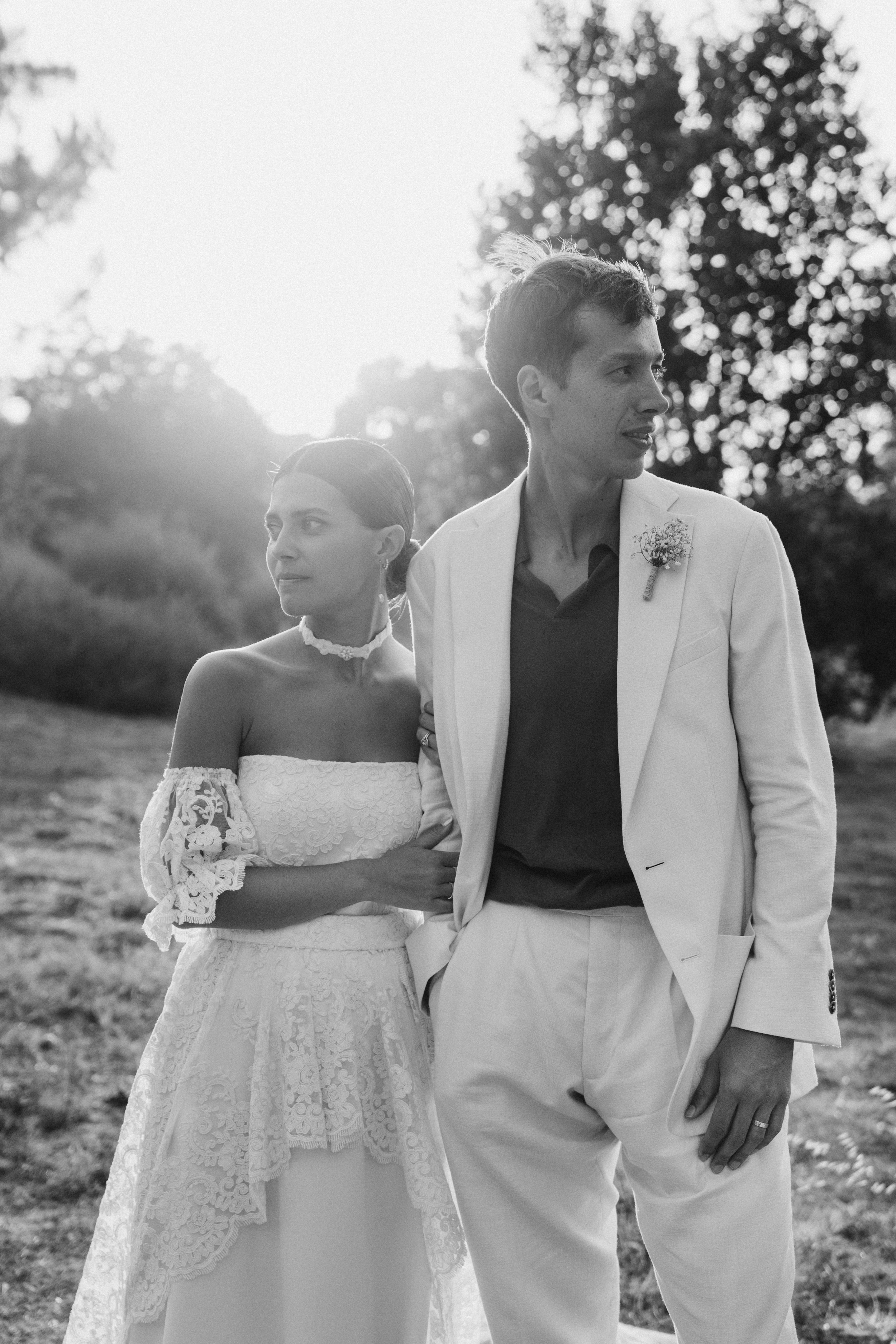 A black and white photo of a couple outdoors, dressed in wedding attire, with the sun setting behind them and trees in the background.